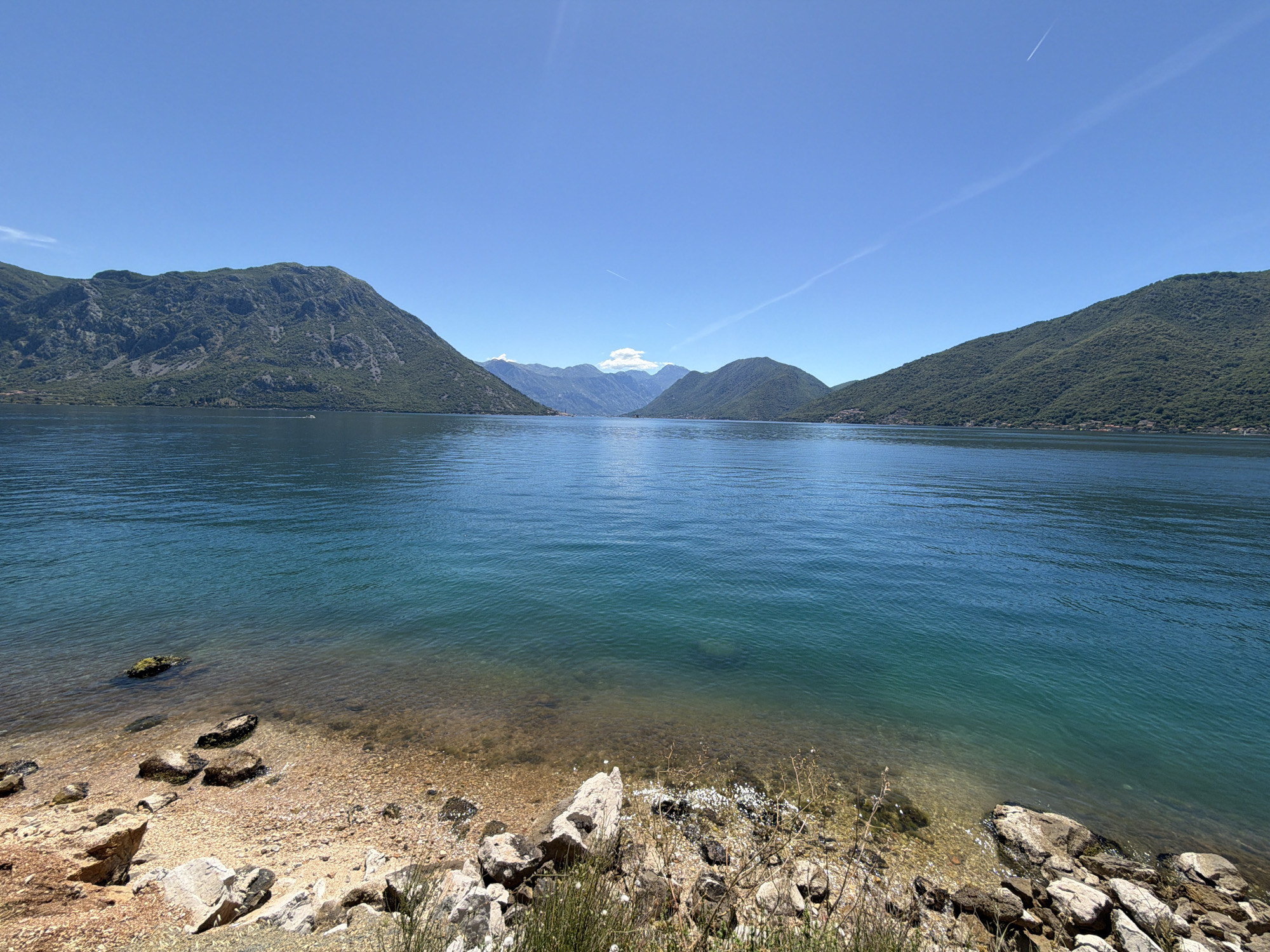 A wide shot of the Bay of Kotor on a clear, sunny day shows the tranquil turquoise water lapping against a rocky shore in the foreground. The water is clear enough to see the pebbles on the seabed near the shore. Lush green mountains rise steeply on either side of the bay, their slopes dotted with trees and, in the distance, a few scattered buildings. The sky is a vibrant blue with a few wispy contrails from airplanes. The scene evokes a sense of peace and natural beauty, with the mountains reflecting in the calm waters. This view is representative of the scenery found in Lipci, Montenegro, a small settlement located on the shores of the bay.