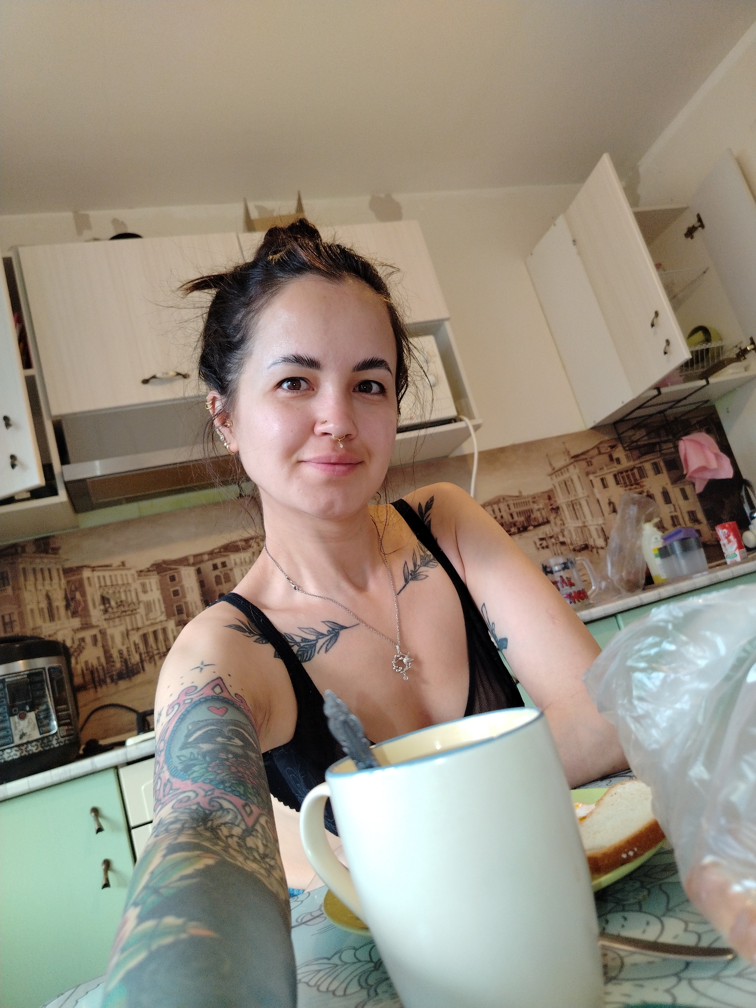 A woman with dark hair pulled back into a messy bun is sitting at a table in a kitchen, likely in Noviy, Russia, given the prompt's mention of the city. She has a nose ring, multiple earrings, and several tattoos, including a full sleeve tattoo on her left arm and other tattoos on her chest and upper arm. She wears a black bra-like top. In front of her is a large white mug with a blue rim, a spoon inside, and a piece of bread on a plate. A plastic bag sits next to the plate. The kitchen has white cabinets, green drawers, and a backsplash depicting a cityscape. An instant pot is visible on the counter. One cabinet door is open.