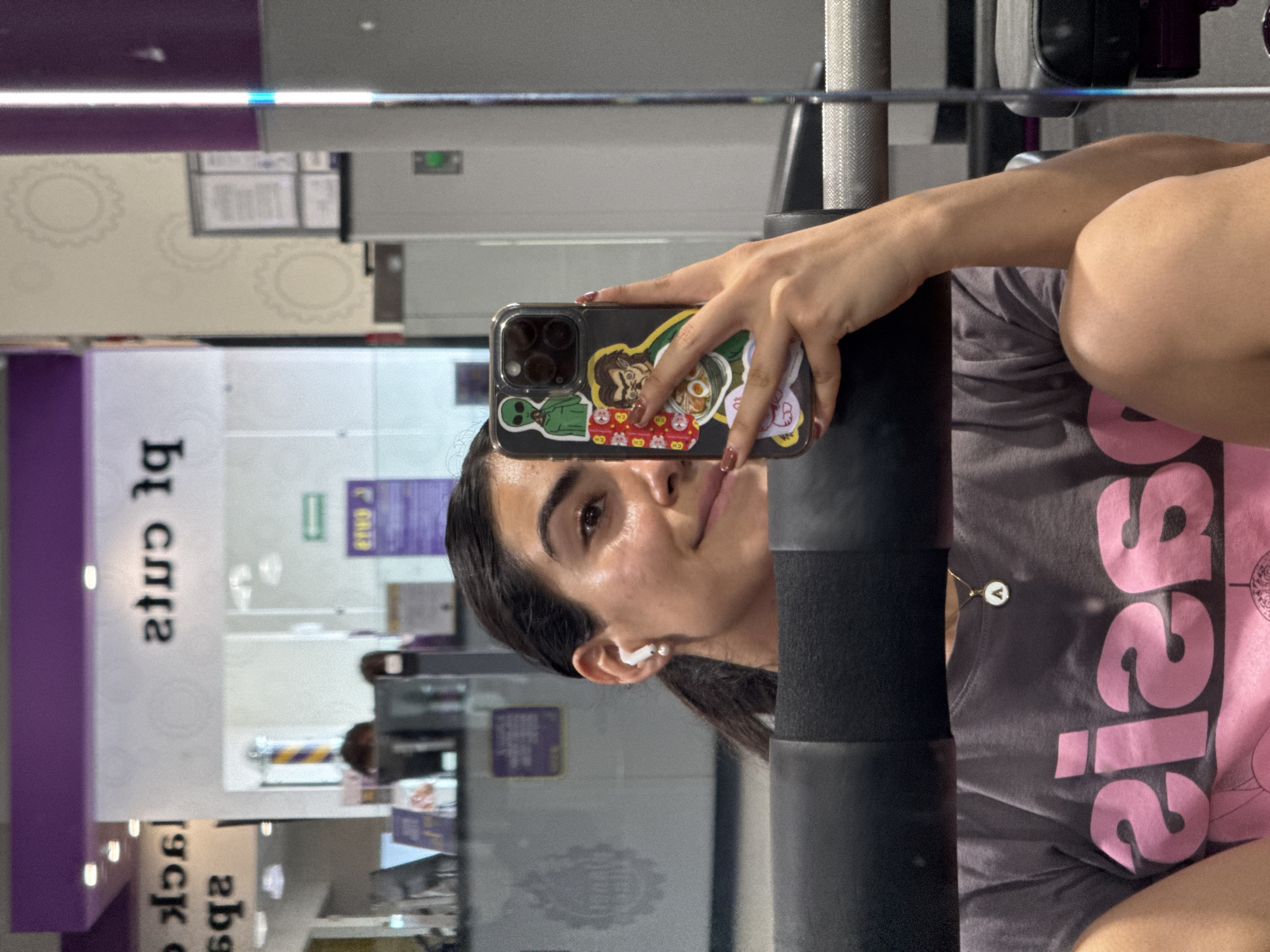 A selfie taken in a mirror in what appears to be a Planet Fitness gym. The person taking the photo is a woman with dark hair, wearing an AirPod in her left ear and a gray t-shirt with pink lettering. Her phone has stickers on it and is covering part of her face. Gym equipment is visible in the background, along with reflections of the gym's interior, which includes purple and white walls and various posters. The gym is located in Puebla, Mexico.