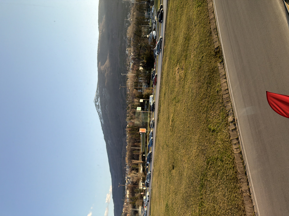 The image depicts a panoramic view of an urban landscape with a prominent natural backdrop under a clear blue sky. In the far background, Vitosha Mountain rises, its highest peak partially covered with snow, while the lower slopes are densely forested with predominantly bare or dark evergreen trees. Below the mountain, a sprawling urban area with numerous buildings, consistent with the city of Sofia, Bulgaria, is visible, interspersed with trees and streetlights.
In the midground, a multi-lane asphalt road carries a line of vehicular traffic, consisting of numerous cars, including sedans, SUVs, and vans, appearing to be in motion or experiencing congestion. Streetlights are regularly spaced along this road. An orange commercial sign and other less legible signs are visible near the roadside.
The foreground features a wide, gently sloping embankment covered with dry, yellowish-green grass, indicating early spring or late autumn. Below this grassy area, the edge of another asphalt road is visible, running horizontally across the bottom of the frame. A distinct red, curved object, possibly part of a vehicle's interior or a reflection, is present in the lower right corner of the image, partially obscuring the foreground road. The scene is illuminated by natural daylight, casting a clear, bright light across the landscape. The photograph is taken from an elevated vantage point, looking down towards the road and across the city towards the mountain.