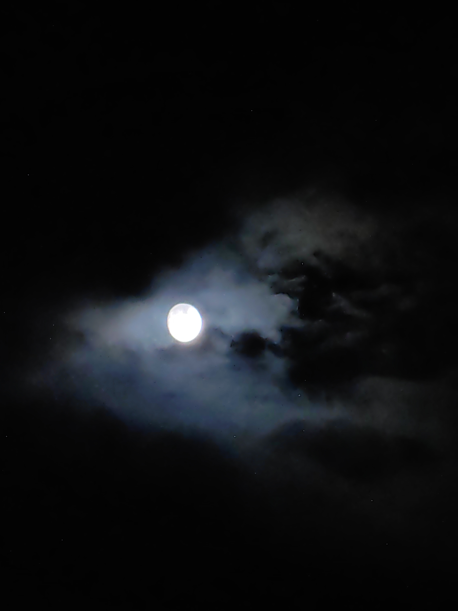 The image shows a full moon partially obscured by dark, wispy clouds against a black night sky. The moon is bright and luminous, casting a soft glow that illuminates the edges of the clouds. A few faint stars are visible in the background. The scene depicts a natural, nocturnal environment, likely captured outdoors in an area with minimal light pollution. There is no indication of Palchan, India, or any urban setting.