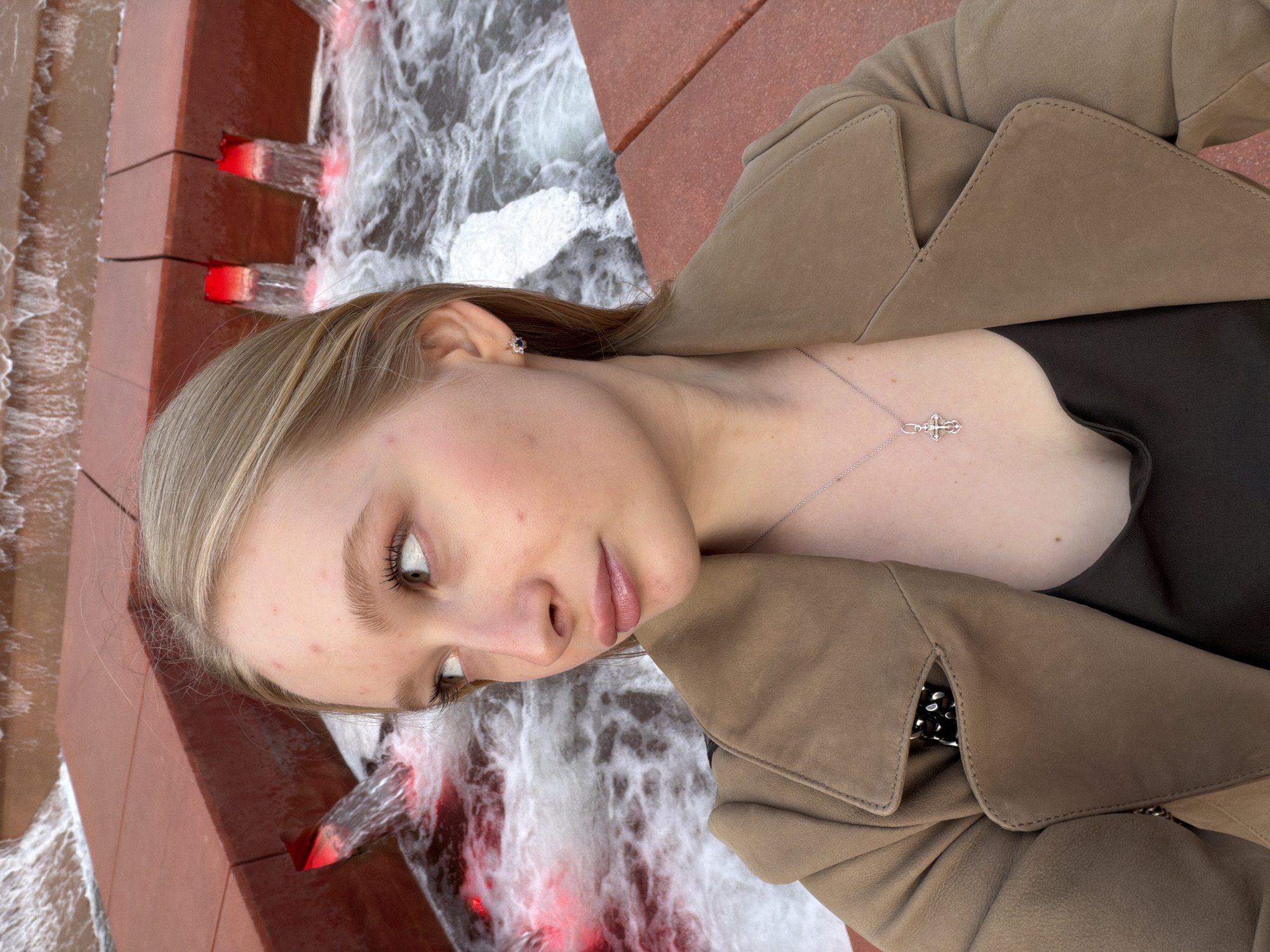 A young woman is posing next to a waterfall feature in the city of Krasnodar, Russia. The waterfall appears to be a modern design, with water cascading over red-toned brick or stone tiers. The water itself is foamy and turbulent. The woman has light blonde hair, fair skin, and is wearing a tan-colored trench coat over a dark top. She is also wearing a necklace with a cross pendant and a flower-shaped earring. She is looking slightly down and towards the camera with a neutral expression.
