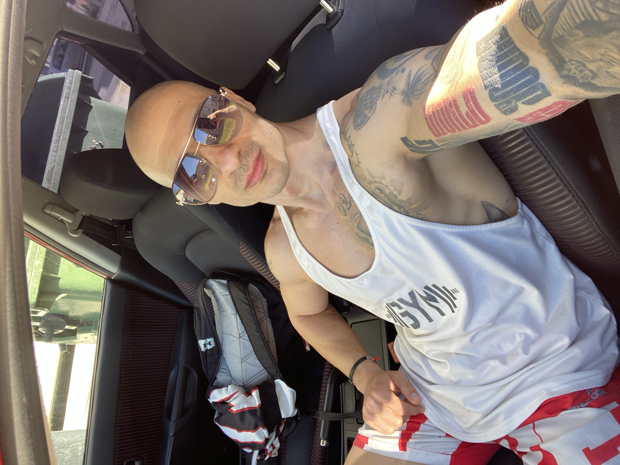 The photograph depicts a man seated inside a car. He is bald, wearing sunglasses, a white tank top with "GYM" printed on it, and red and white shorts. He has several tattoos on his arms and chest. A backpack and a garment with black and red stripes are on the seat next to him. The car's interior is visible, including the black seats. Reflections can be seen in his sunglasses. Based on the reflections in the sunglasses, the location is Pleven, Bulgaria, evidenced by the unique architectural features of buildings visible in the reflections, specifically the distinct balconies and window structures characteristic of Pleven's architecture. The man is likely taking a selfie, holding his arm out to capture the image.