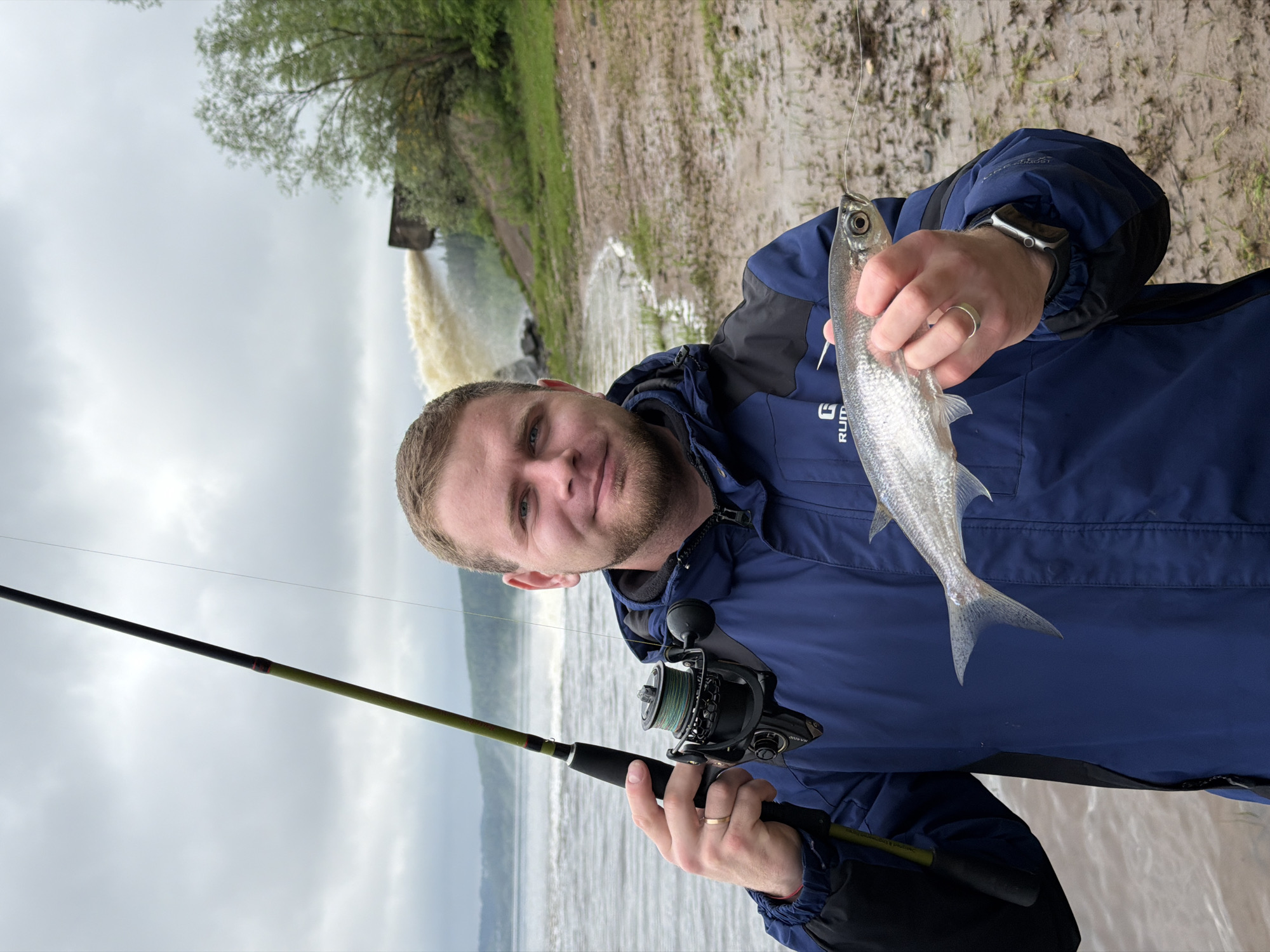 A man stands outdoors, holding a fishing rod in his left hand and a fish in his right. He wears a blue jacket over a black hooded shirt, and a ring is visible on his left ring finger. He smiles at the camera. The fishing rod is black and green with a reel. The fish is silver and appears to be freshly caught. In the background, a waterfall or dam structure empties into a body of water. Trees line the bank behind the man and the water. The sky is overcast. The location appears to be in the city of Ольховка, Russia.