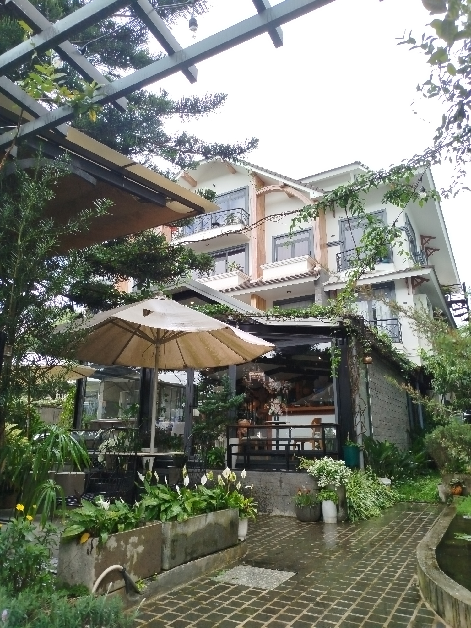 This image depicts the exterior of "MC House," a coffee shop and lodging located in Da Lat, Vietnam. The building is a multi-story structure with white walls and grey-trimmed windows. A dark metal pergola extends over an outdoor seating area where tables and chairs are visible. A beige umbrella provides shade over one of the tables. Lush greenery, including trees, potted plants, and flowering shrubs, surrounds the establishment, creating a garden-like atmosphere. The ground is paved with dark square tiles, and a narrow concrete pathway leads to a small pond. The sky is overcast, suggesting a cloudy day. The scene appears calm and serene, with no people visible.