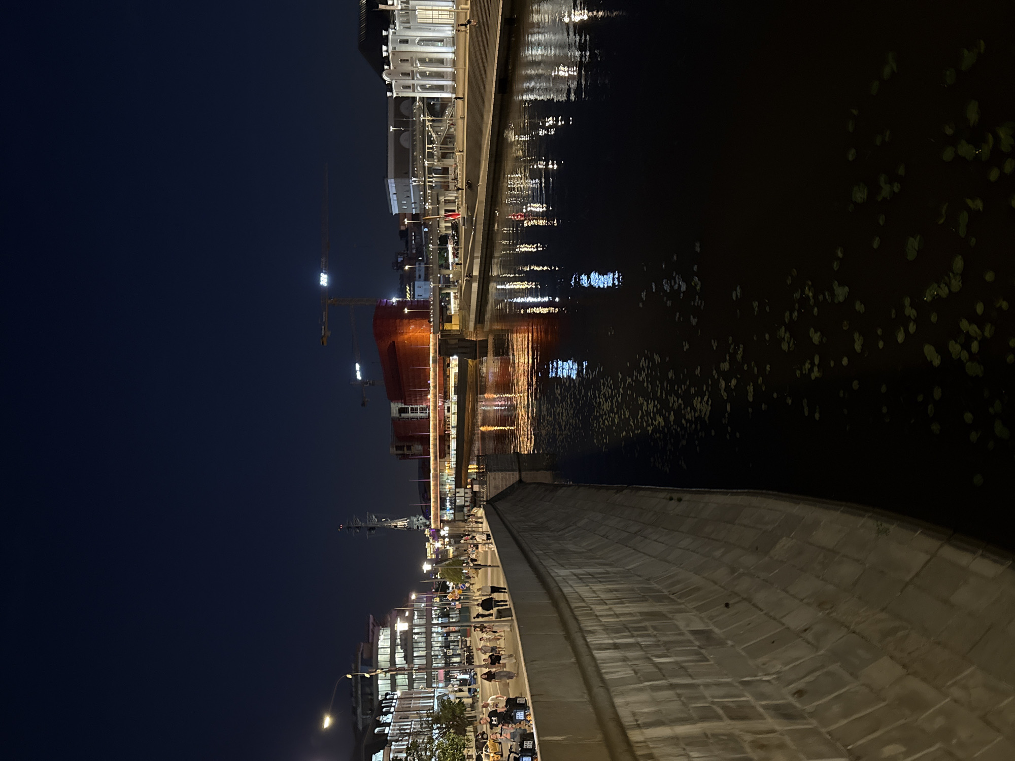 This nighttime image captures an urban scene along a canal or river embankment in Moscow, Russia. The perspective is from an elevated position on a curved, stone-block embankment, looking across a body of water towards the opposite bank.
In the foreground and mid-ground on the left, a sturdy, light-colored block-work wall curves gently along the water's edge. Above this wall is a pedestrian walkway, well-lit by streetlights, where approximately two dozen individuals are visible, walking, standing, and conversing. Beyond the walkway, a street is discernible with several parked cars and a yellow taxi. Buildings with numerous illuminated windows line this street, and a tall, dark, mast-like structure with visible rigging rises above them in the distance.
The central and right portions of the image are dominated by the dark, placid water, which vividly reflects the city lights in vertical streaks of yellow, white, and red. Scattered across the water's surface are numerous small, dark green, rounded objects resembling lily pads or aquatic vegetation.
In the mid-distance, a multi-tiered bridge spans the water, featuring a lower road deck and an upper structure, with elements illuminated in orange. On the far bank, a series of terraced steps descends towards the water, where several people are present. Further back, a prominent, brightly illuminated white building with classical columns and large windows stands out. To its left, a more modern, lighter-colored building is visible.
The background reveals a deep, dark blue-black sky. Two large construction cranes with illuminated booms are visible in the distance, one bearing the prominent white letters "ПИК." Another building under construction, draped in red netting, is also discernable.
The scene depicts a lively urban environment after dark, characterized by architectural diversity ranging from classical to modern, active pedestrian areas, and ongoing construction. The location is in the vicinity of the Vodootvodny Canal in Moscow, Russia, likely viewed from either the Bolotnaya or Kadashevskaya Embankment, looking towards the Bersenevskaya Embankment and the "Krasny Oktyabr" (Red October) complex.