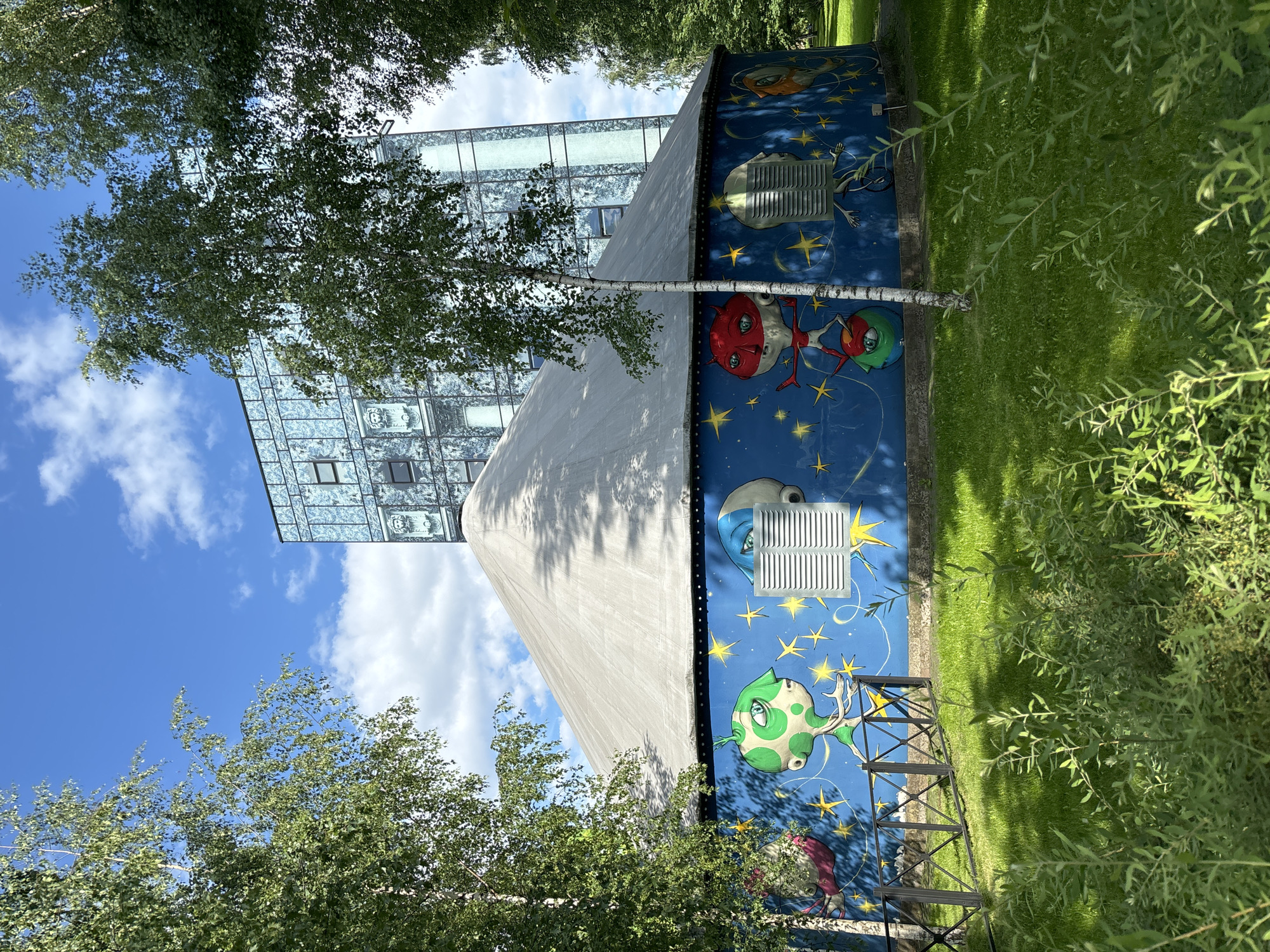 The image depicts a cylindrical building with a conical roof, adorned with a vibrant mural on its blue facade. The mural features whimsical, alien-like characters with large eyes, set against a starry background. Two white, louvered vents are visible on the mural. A slim, light-colored tree stands in front of the building, partially obscuring the mural. Behind this structure, a modern, multi-story building with a glass facade is visible. The scene is set in a green, grassy area with lush foliage in the foreground and trees framing the shot. The sky is blue with scattered white clouds. This building is the Neva Color (Neva Tsvet) or Children's Art Center, located at 48 Nevsky Prospekt, Saint Petersburg, Russia.
