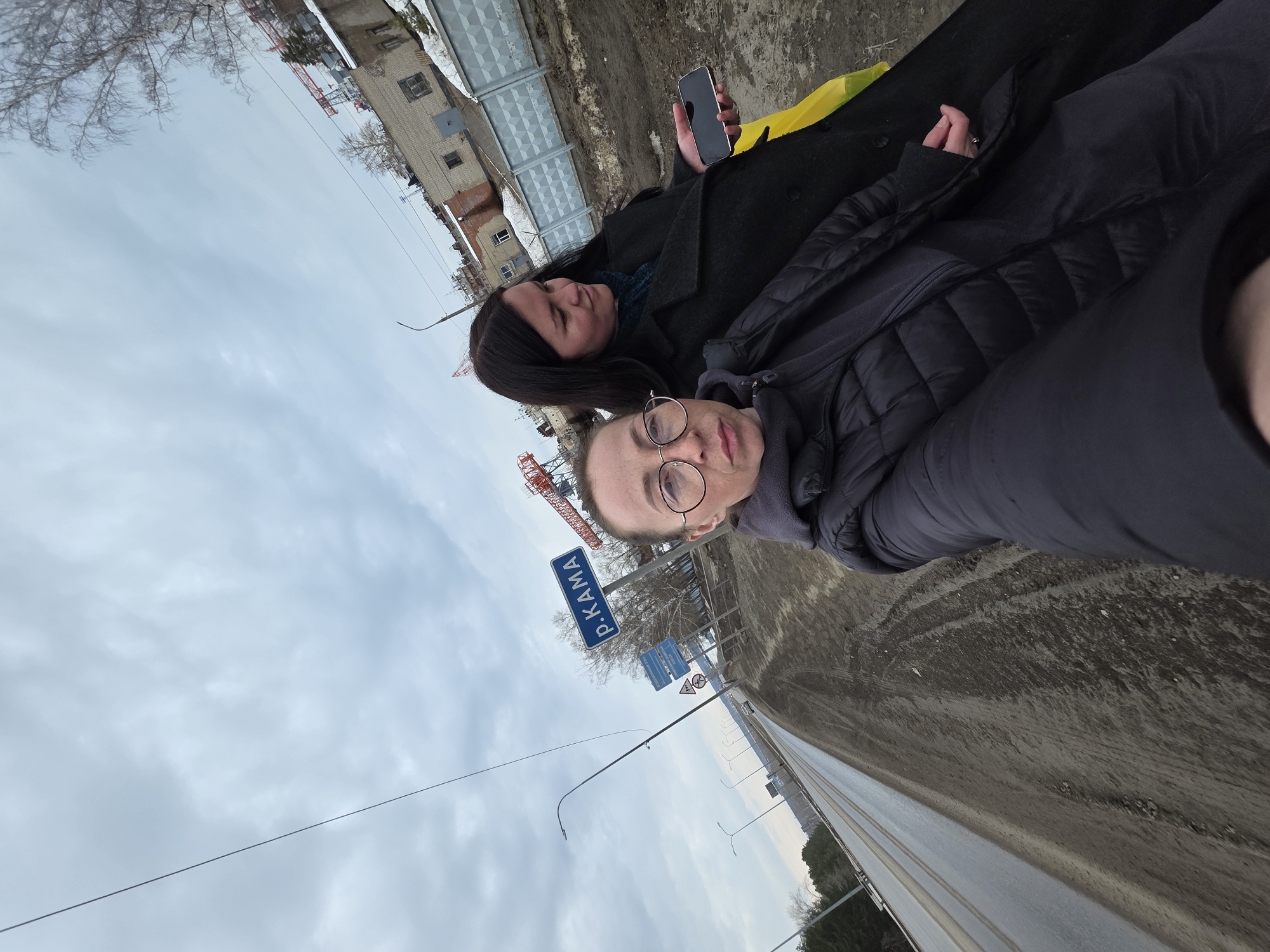 Two women are in an outdoor selfie along a road with a sign reading "Р.КАМА" in Cyrillic letters, indicating they are near the Kama River. The woman in the foreground is wearing glasses and a dark jacket. The woman behind her is holding a phone. The background includes buildings, a construction crane, and a cloudy sky. This is likely in or near Chaikovsky, Russia, given the location near the Kama River.