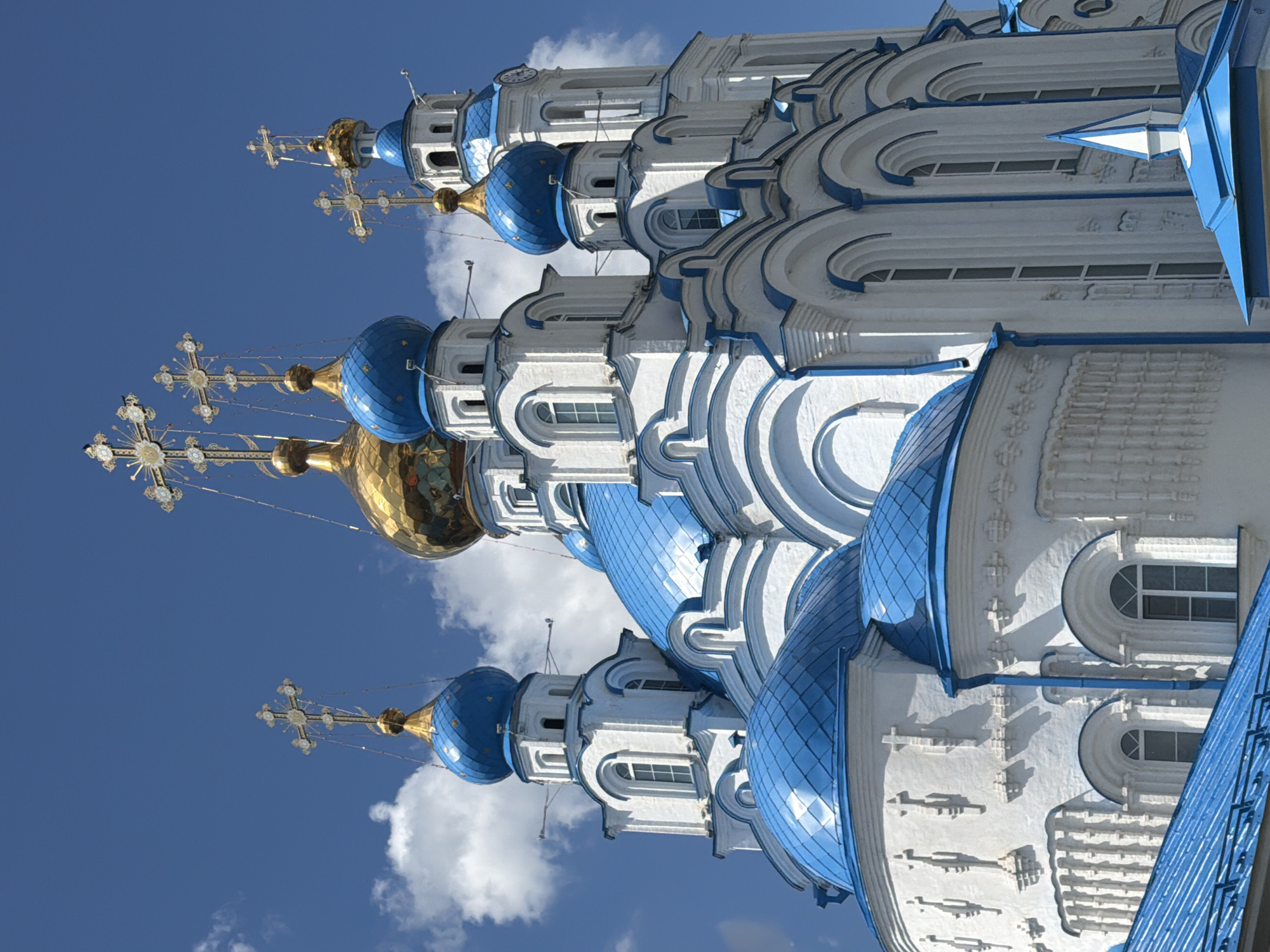A low-angle shot captures the white and blue domed spires of an Orthodox Christian church against a bright blue sky dotted with white clouds. The church features multiple domes of varying sizes, most of which are a vibrant blue and topped with ornate golden crosses. One larger dome, centrally positioned and slightly behind others, is entirely golden. The white facade of the church is adorned with architectural details, including arched windows and decorative elements. The image appears to be a photograph of a religious building, likely a cathedral or a large church, emphasizing its architectural grandeur and its spiritual symbolism.