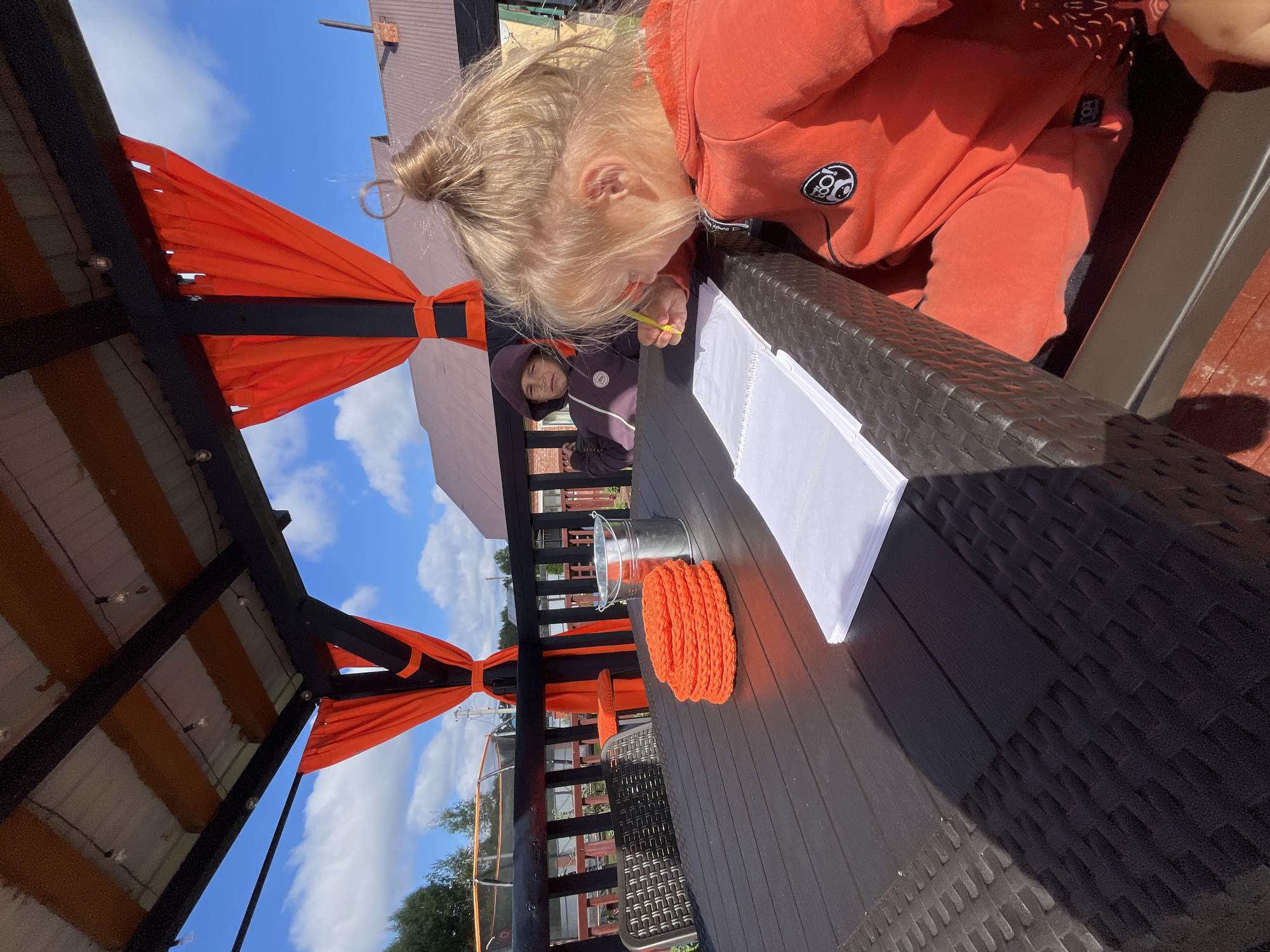An outdoor shot captures two children sitting at a dark brown table under a covered patio with orange curtains. The girl in the foreground, facing right, has blonde hair tied in a bun and is wearing an orange hoodie. She is drawing in a white notepad with a yellow pencil. Behind her, a boy in a purple hoodie is looking towards the camera with a neutral expression. On the table, to the left of the notepad, is a stack of orange crocheted coasters and a metal bucket. The patio structure is visible with black beams and a corrugated metal roof. In the background, trees, houses, and a trampoline are discernible under a blue sky with scattered clouds. The location is identified as Chaikovsky, Russia.