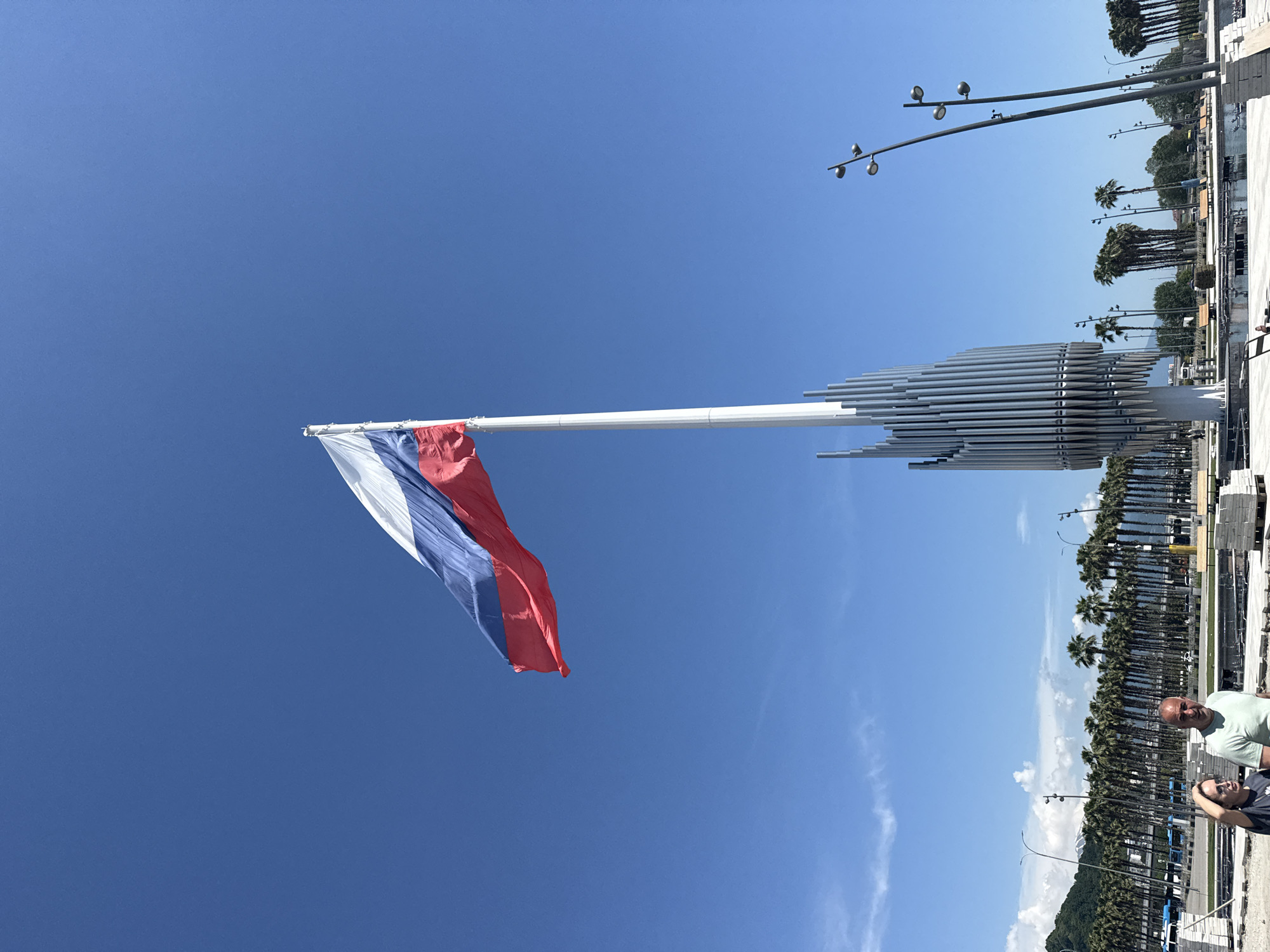 The image captures a wide shot of the Sirius Olympic Park in Sirius, Russia, on a bright, clear day. Dominating the upper center is a large Russian flag, horizontally striped white, blue, and red from top to bottom, unfurling to the right against a brilliant azure sky. It flies from a very tall, slender white flagpole.
To the right and below the flagpole's base stands a distinctive, modern monument composed of numerous vertical, silvery metallic tubes of varying heights, tapering towards the top. This structure resembles a large organ pipe assembly and has a somewhat architectural or sculptural quality.
In the bottom left foreground, two individuals are visible. A woman with dark hair, wearing sunglasses and a dark t-shirt, looks towards the left with her left hand near her head. Beside her, a man with a shaved head and a light green t-shirt looks directly towards the viewer with a slight smile. They appear to be walking in an area that seems to be a public square or promenade, partially under construction or renovation, with stacks of paving stones or other building materials visible on the ground.
The midground features a long row of uniform palm trees, lining what appears to be a wide road or public space. Beyond the palm trees, various structures and vehicles are faintly visible, including what look like blue buses. In the far distance, low mountains are visible under a horizon line that shows faint wisps of white clouds. Several tall, dark streetlights with multiple lamp heads are scattered across the scene, particularly on the right side. The overall impression is one of a vast, sunny, and somewhat open public area.