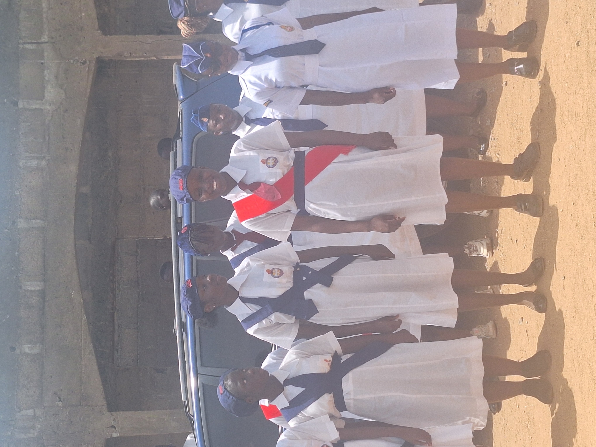 A group of young girls in white uniforms with blue accents stand in a line outdoors in Sabon Tasha, Nigeria. The girls wear matching blue caps with small red emblems, white dresses, and dark shoes. Some wear blue ties or sashes, while one wears a red sash. They are lined up next to a light blue vehicle. A grey concrete building is visible in the background. The ground is sandy and sunny.