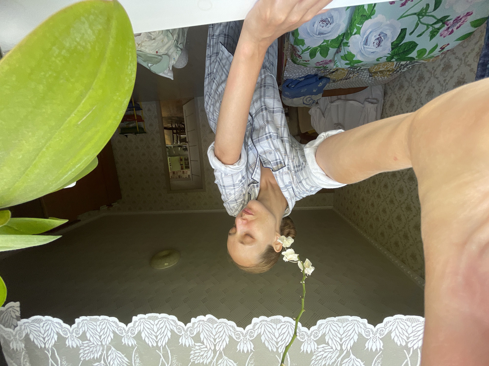 A woman with light brown hair pulled back into a bun and white flowers tucked behind her right ear is seen from the chest up, looking down with her eyes closed. She is wearing a light blue and white plaid pajama top with rolled-up sleeves. Her left arm and hand are extended towards the camera, obscuring a portion of the frame. The background appears to be an interior residential space, possibly a bedroom, with floral patterned wallpaper. A bed with blue and white floral patterned bedding is visible to the left. A doorway leading to another room is visible in the background. The scene is illuminated by natural light, suggesting it is daytime. The provided metadata indicates the location is Sharkan, Russia.