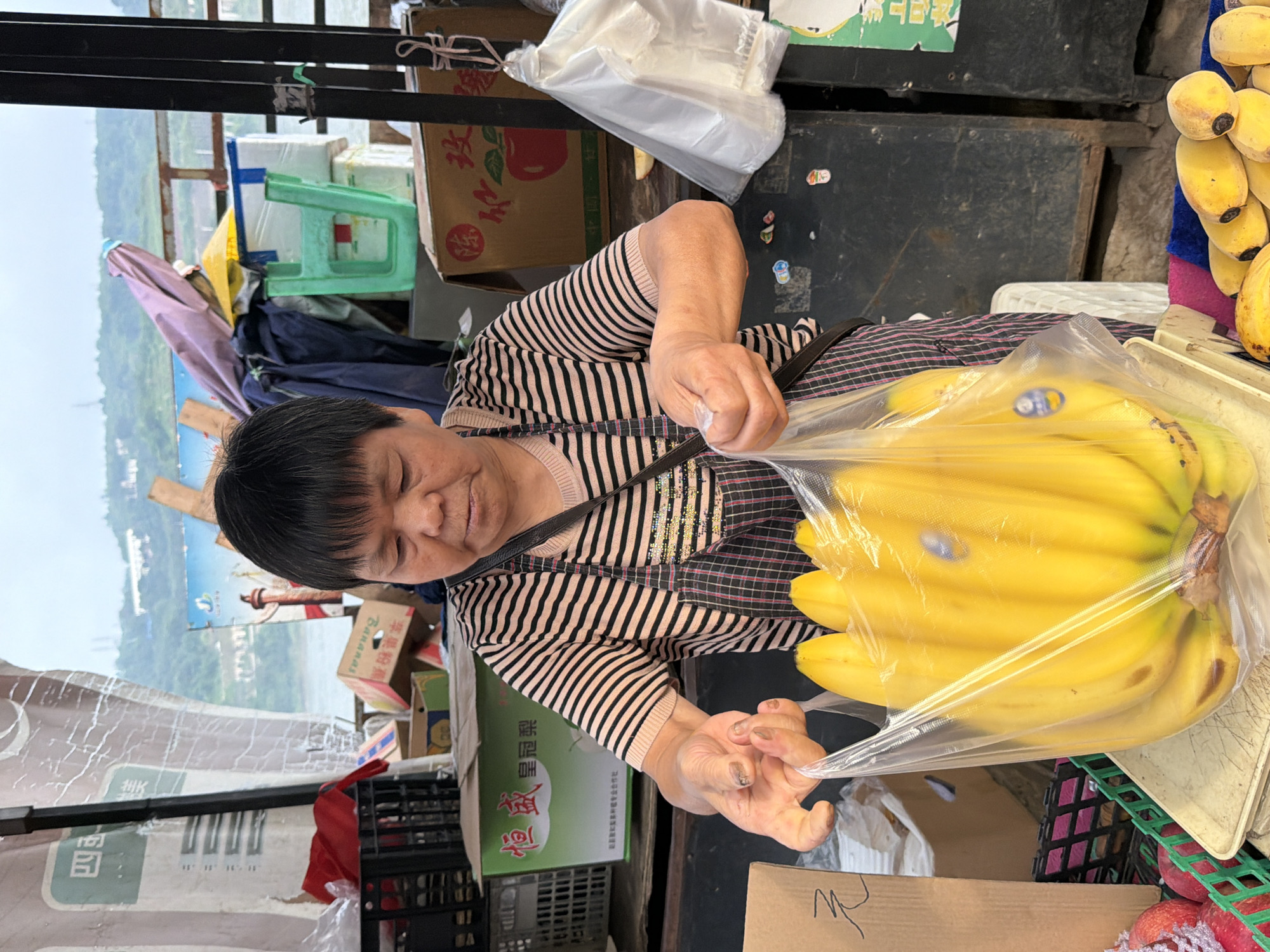 A woman with short black hair is standing behind a market stall, holding a clear plastic bag filled with bright yellow bananas. She is wearing a pink and black striped shirt and a black and red plaid apron. Her hands are inside the bag, adjusting the bananas. The market stall is cluttered with various items, including cardboard boxes, plastic crates, and other fruits like apples. In the background, there are more boxes, a green plastic basket, and a body of water with hills beyond. The overall scene suggests a bustling outdoor market. The woman is likely a vendor selling produce in Jiangjin City, China.