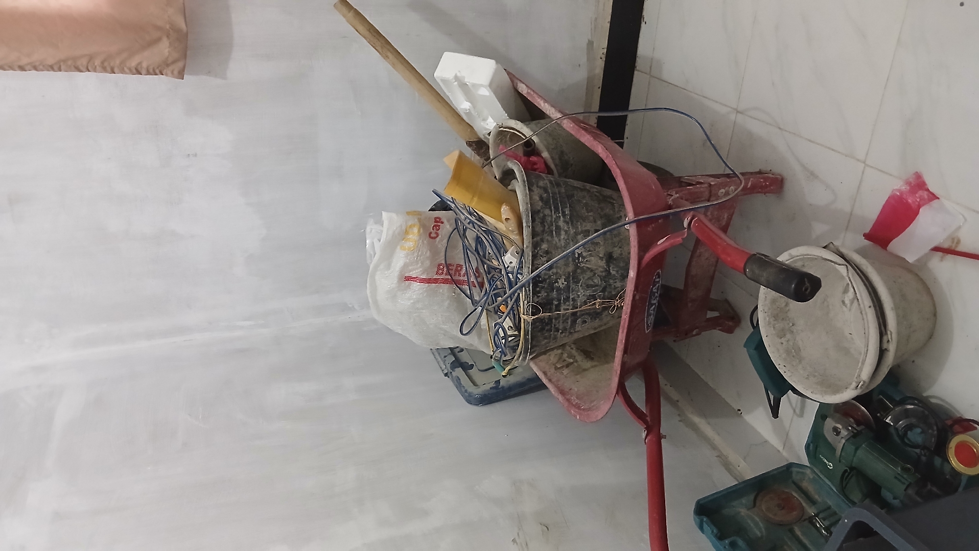 The image depicts a construction site or storage area, likely in Klambir Lima Kebun, Indonesia. A red wheelbarrow is loaded with various items, including a large white plastic bag, several buckets, and a tangle of blue electrical cords. One bucket appears to be made of black plastic, while another, placed in front of the wheelbarrow, is a lighter color and filled with a white powdery substance. To the left of the wheelbarrow, a green power tool, possibly an angle grinder, is housed in a teal and black case. Nearby, a small Indonesian flag, red and white, lies on the floor. The background consists of a white wall that appears to be unfinished or under renovation, with a segment of a beige curtain visible in the upper right corner. The floor is tiled with light-colored squares. The scene is static, with no people or overt actions taking place.