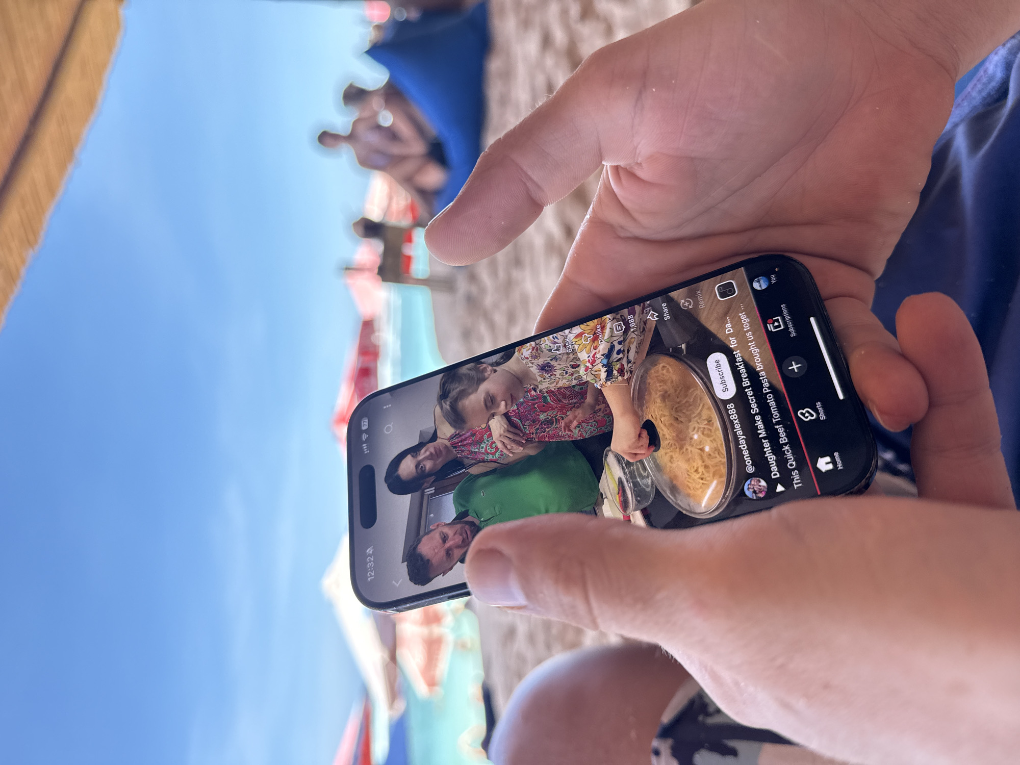Here is a forensic description of the image:
A person is holding a smartphone displaying a YouTube Shorts video featuring a family—a father, mother, and child. The video is titled "Daughter Make Secret Breakfast for Da..." by @onedayalex888, showcasing a quick beef tomato pasta recipe. The phone's screen displays the YouTube interface with options like "Dislike," "Share," "Remix," "Shorts," and "Home." The phone indicates it is 12:32 and has full signal strength.
The setting appears to be outdoors, possibly a beach, as suggested by the sand-like particles on the person's hands, the blue sky, and out-of-focus figures in the background. These figures include individuals sitting under what seems to be a beach umbrella. The person holding the phone is wearing a blue shirt or shorts. The overall environment suggests a sunny, outdoor leisure setting, consistent with a location like Kuta Selatan, Indonesia.