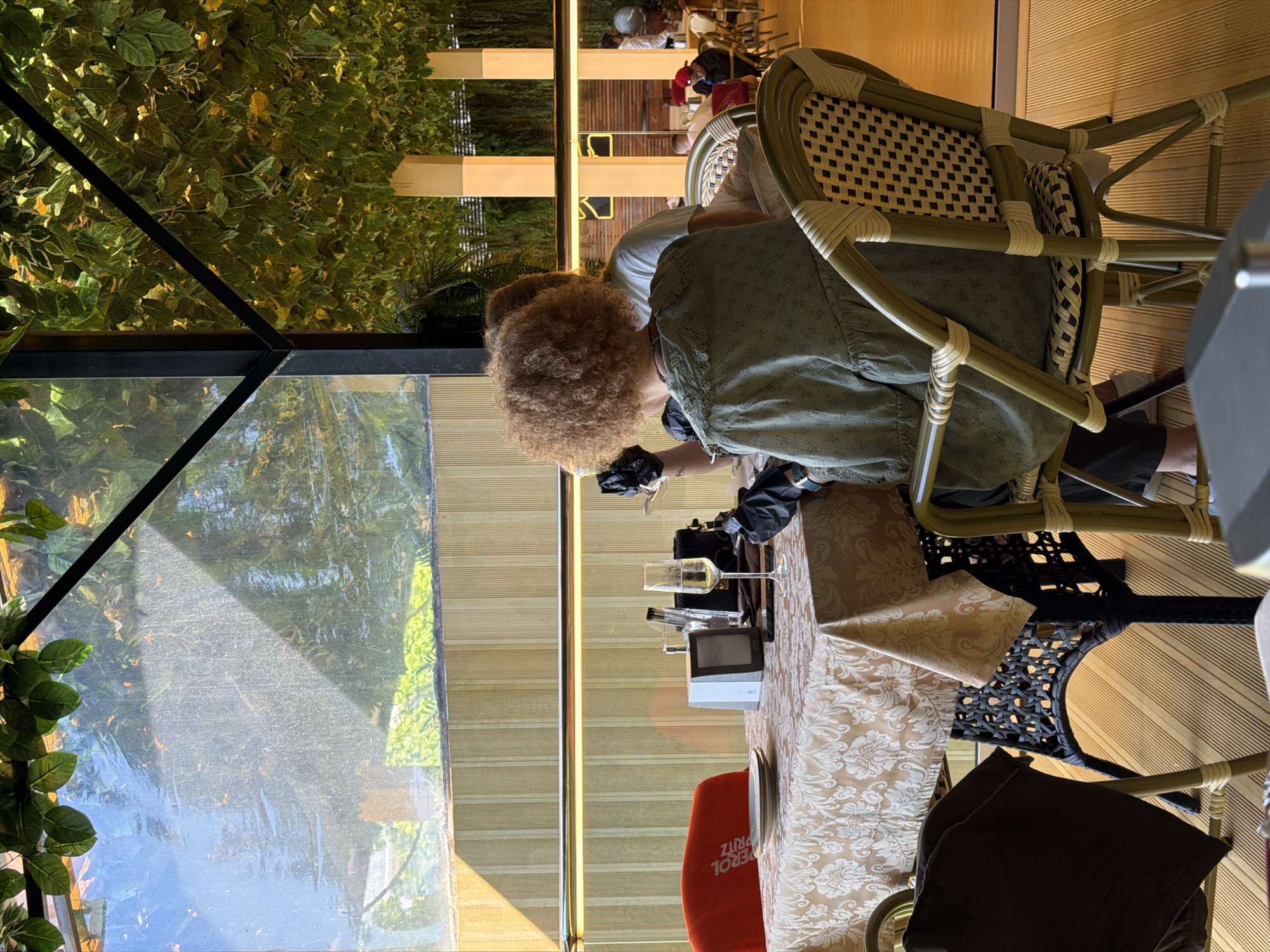 The image depicts a restaurant interior, possibly in Svetlogorsk, Russia, with a focus on two individuals seated at a table. The table is covered with a patterned tablecloth and set with plates, a wine glass filled with a bubbly beverage, and a small silver device. One individual, facing away from the camera, has curly auburn hair and is wearing a light green patterned blouse. They are wearing black gloves and appear to be holding something near their face. The second individual, partially visible, is wearing a white t-shirt. The restaurant features a backdrop of lush green foliage, some of which hangs from the ceiling, creating a covered outdoor or conservatory-like ambiance. Through a transparent screen in the background, glimpses of other patrons and restaurant elements are visible. The lighting suggests it is daytime, with sunlight casting shadows. The flooring is light-colored wood.
