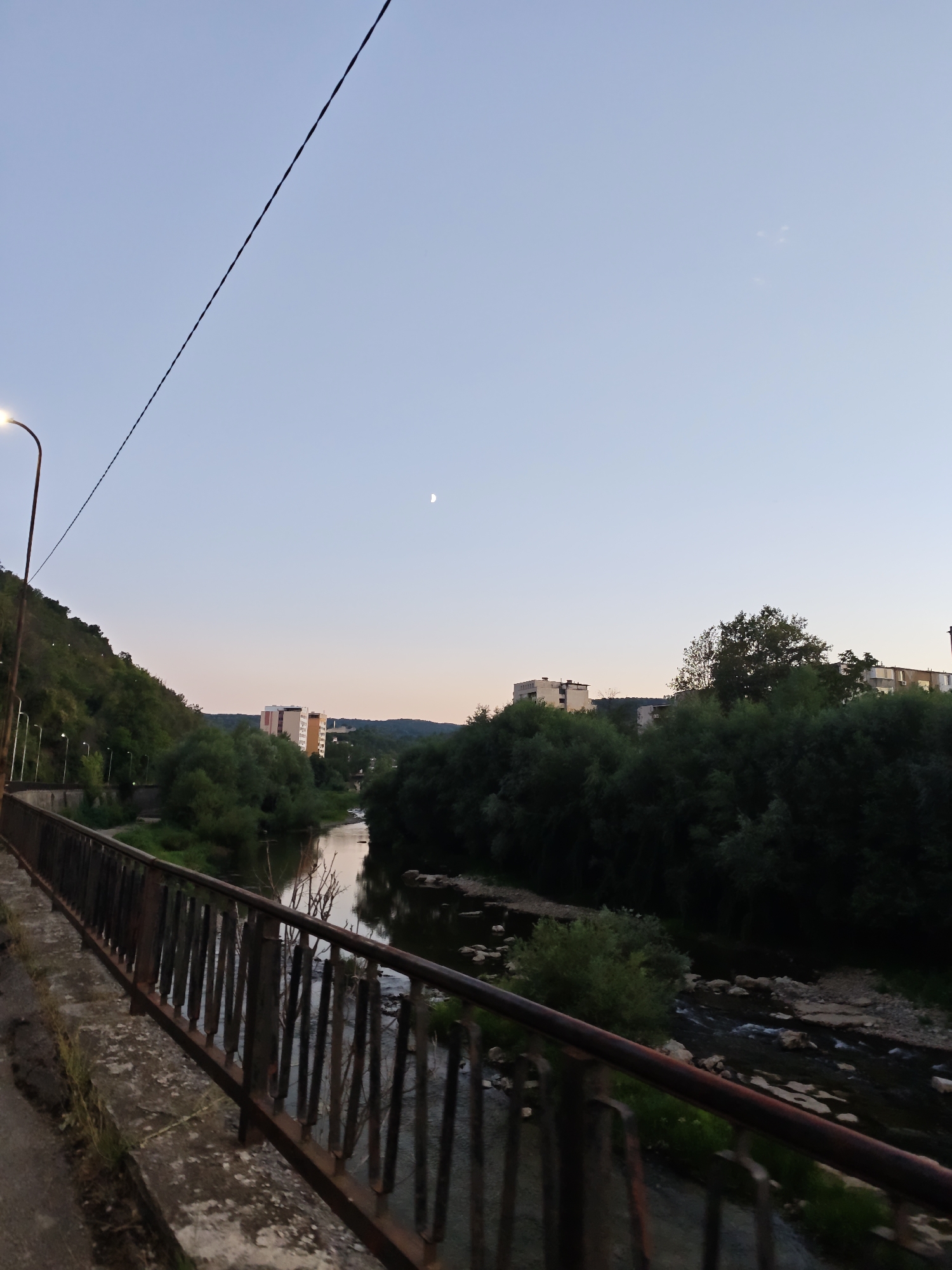 The image captures a twilight view of the Osam River in Lovech, Bulgaria. In the foreground, a rusty metal railing lines a concrete walkway. The river flows gently, reflecting the pale sky and surrounding greenery. On the left bank, a steep, tree-covered hillside is illuminated by streetlights. Across the river, apartment buildings and dense foliage are visible. The moon is a faint crescent in the clear sky. A power line stretches diagonally across the upper portion of the frame.