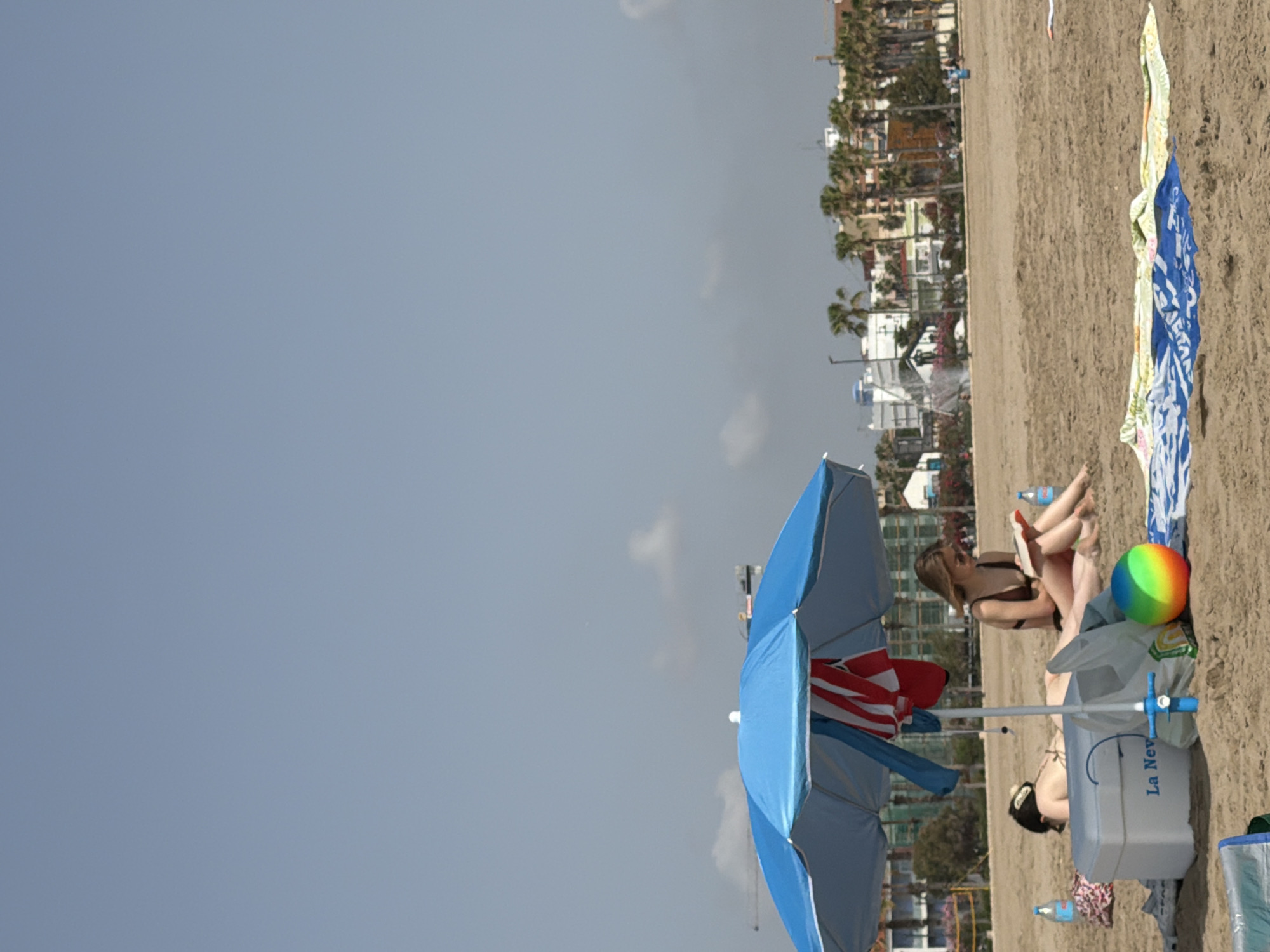 In an outdoor scene of a sandy beach, under a clear sky, a blue beach umbrella shades two individuals. The person on the left, partially visible, lies on their stomach. The person on the right, a woman wearing a brown bikini, sits up and reads a book. A cooler is placed on the sand near the umbrella, along with a rainbow-colored ball and other beach items. In the background, palm trees and buildings can be seen, suggesting an urban coastal setting. The overall atmosphere is one of relaxation and leisure.