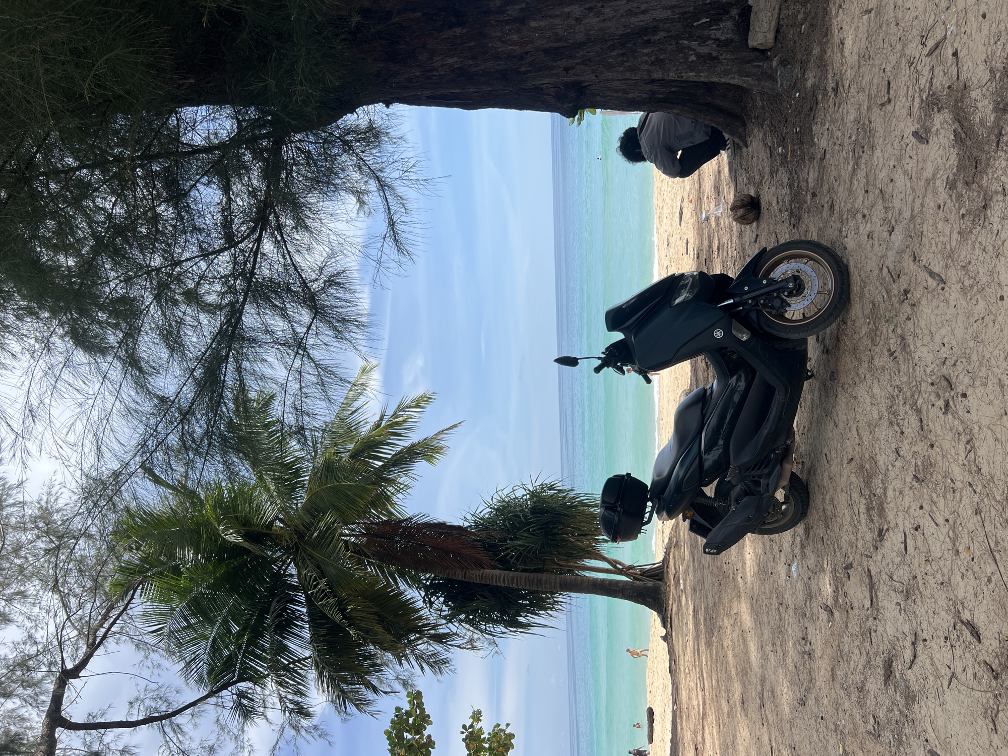 A black scooter is parked on a sandy beach under the shade of trees. The scooter is facing left, with its front wheel turned slightly inward. Behind the scooter, a person is crouched down, facing away from the camera, near the base of a large tree. The beach extends to the right, meeting the turquoise ocean. Several palm trees are visible further down the beach, and a few people can be seen in the shallow water. The sky is blue with scattered clouds. The scene appears to be in Choeng Thale, Thailand.