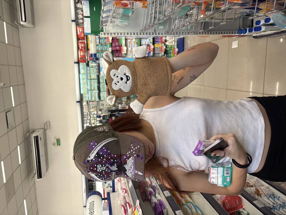 A woman with auburn hair wears a white tank top, black pants, and a decorated baseball cap in a store. A brown monkey hand puppet sits on her shoulder. She holds a cell phone and several small packages in her hand. Shelves filled with various products are visible in the background. The scene is brightly lit, suggesting a commercial environment like a pharmacy or convenience store.