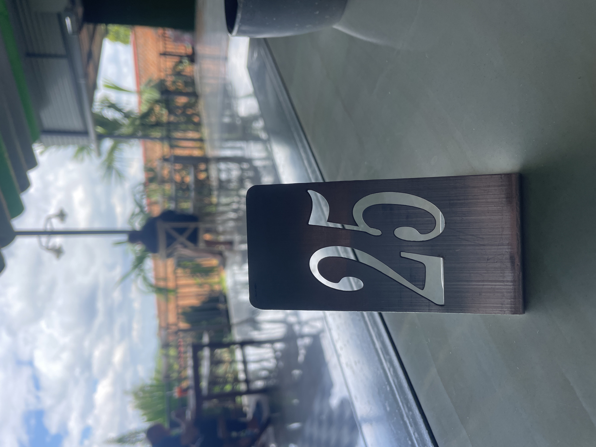 The image presents a detailed view of a cafe or restaurant setting in Chư Sê, Vietnam, during daytime, with a sharp focus on a table number. The primary subject is a dark, brushed metallic table number "25," prominently displayed in the lower-middle frame. The numbers are cut out, revealing a lighter, reflective material underneath. The table number stands upright on a highly reflective, light-colored surface, possibly a polished table or floor, which shows blurred reflections of the bright sky and overhead structures.
To the right of the table number, a portion of a grey, speckled ceramic mug or cup is partially visible, out of focus. The overhead area features a green, scalloped awning or roof structure and dark, possibly metallic, support beams.
The background, largely out of focus, reveals an outdoor environment under a bright, partly cloudy sky with scattered white cumulus clouds. Numerous palm trees and other green foliage are visible. Distant red-tiled rooftops, characteristic of local architecture, are apparent, separated by what appears to be a dark fence or railing. On the far left, a severely blurred figure is seated at a table, with what seems to be a dark beverage in a glass visible next to them. A dark, ornate street lamp-style fixture with multiple lamps extends vertically on the right side of the background. The floor in the background also exhibits reflective qualities, suggesting a polished surface or recent moisture.