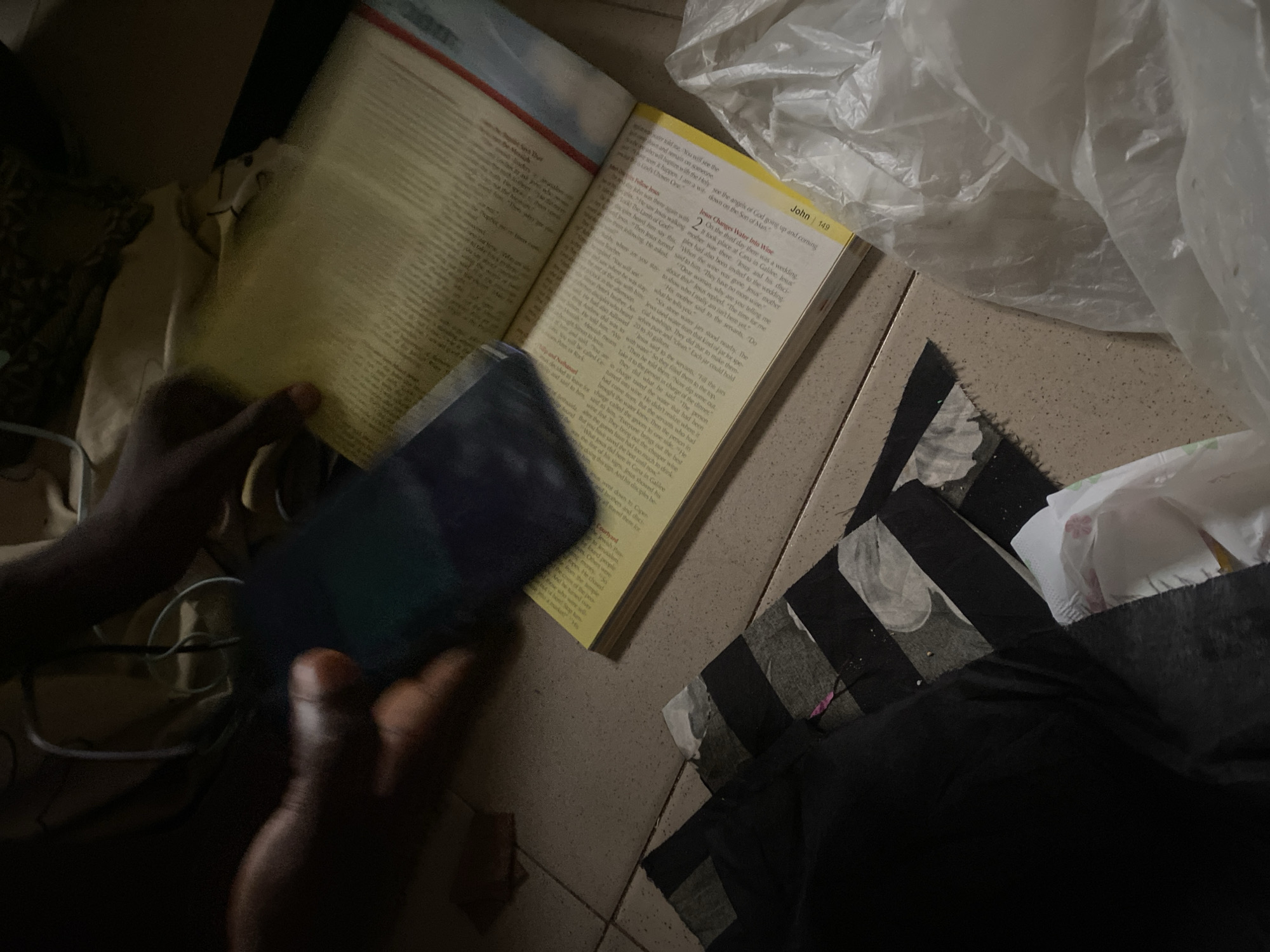 The image shows a close-up of a person's hands holding a smartphone over an open book. The book appears to be a religious text, possibly a Bible, with visible text and page numbers. The scene is dimly lit, suggesting an indoor environment, possibly a home. The floor is tiled. There are some fabric scraps and a plastic bag in the foreground. The image does not provide any information about the location in Jauro Yinu, Nigeria, nor does it contain any details that would indicate such a location.