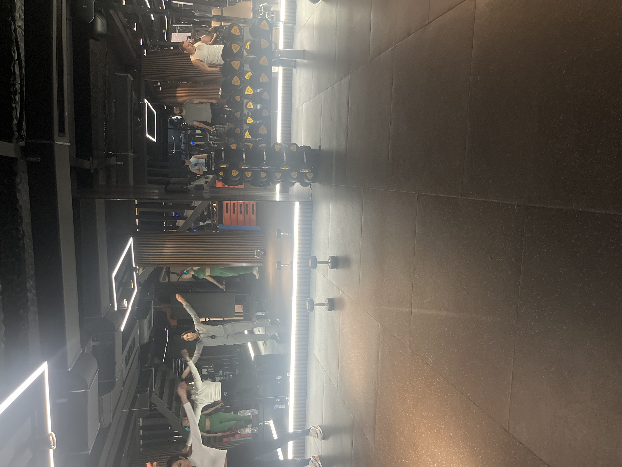 The image shows the interior of a gym, likely located in Sofia, Bulgaria. The gym features rows of weights and exercise equipment, arranged linearly on the left side of the frame. Reflected in a mirrored wall are several people, including individuals exercising and potentially instructors leading a class. The gym has a modern design with dark flooring and ambient lighting. The mirrored wall also reflects the weight equipment and the gym's interior, creating a sense of depth and spaciousness.