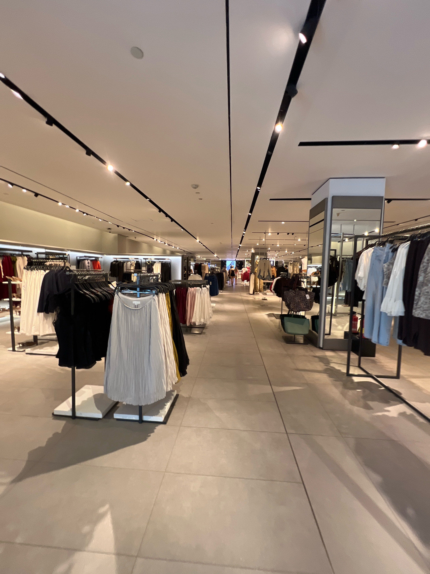 The image depicts the interior of a modern clothing store, likely a boutique or a department store. The lighting is bright and even, illuminating racks of clothing organized by color and style. On the left side of the frame, clothing is displayed on racks with clean, minimalist stands, featuring skirts and dresses in white, black, and light grey. In the center, a large mirror column reflects the clothing displays and the overall store layout. To the right, more racks of clothing are visible, including blouses, dresses, and outerwear in various colors such as blue, white, black, and grey. The flooring is tiled in a neutral, light gray color, and the ceiling is equipped with track lighting. In the distance, blurred figures of customers can be seen browsing the merchandise, suggesting an active retail environment. The overall aesthetic is clean, organized, and upscale, typical of a fashion-focused retail space. Based on the context of Kuta, Indonesia, this scene is likely a fashion store located within a mall or a shopping street in the area.