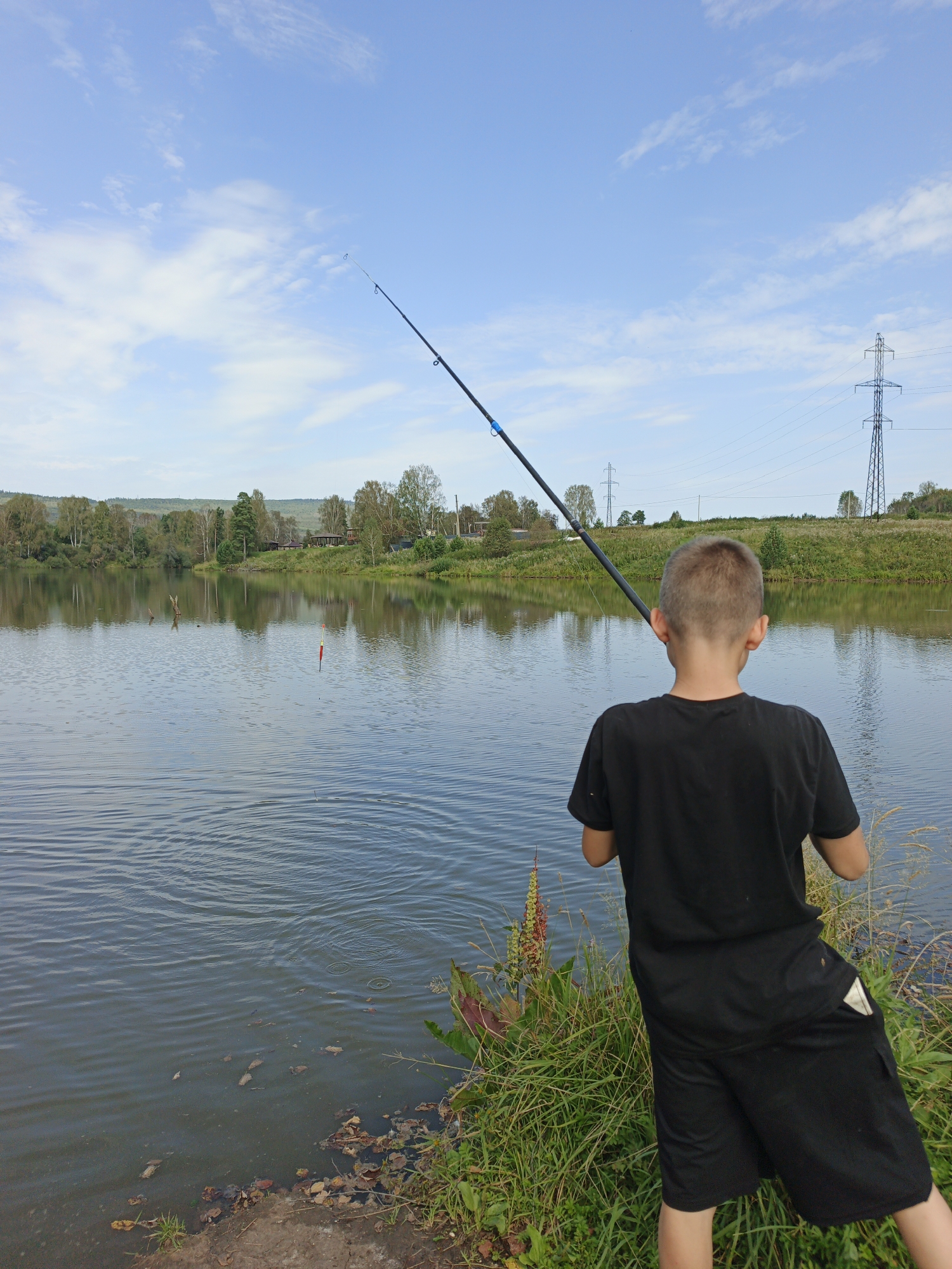 In Urtiushka, Russia, a young boy, facing away from the camera, is fishing on the shore of a calm lake. He is wearing a black t-shirt and black shorts. A fishing rod is held in his hands, with the line extending into the water where a red and white bobber is visible. The background shows a lush green landscape with trees and vegetation, reflecting in the water. Power lines and a tall electrical tower are visible in the distance, indicating the presence of infrastructure near the natural setting. The sky is blue with scattered white clouds. The ground near the boy is covered with grass and fallen leaves.