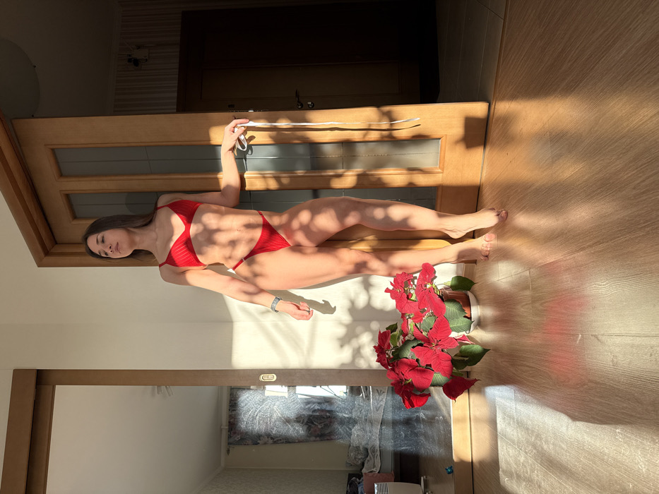 An indoor scene depicts a female individual standing barefoot on a light wooden floor. She wears a two-piece red bikini and has long, dark hair. Her left arm is by her side, adorned with a light-colored bracelet, while her right hand holds a long white strap. She leans against a light-colored wall or door frame, with her head subtly tilted upwards and to the right. Strong natural sunlight illuminates the subject and the immediate foreground, casting distinct dappled shadows of foliage patterns on the wall and floor. To the left, a potted Poinsettia plant with vibrant red bracts rests on the floor in a terracotta-colored pot. In the background, to the left of the individual, a large sliding mirror door reflects an adjacent room containing a white radiator and various household items. To her right, a light wooden hinged door with frosted glass panels is slightly ajar, revealing a darker interior space. The setting is an apartment located in Saint Petersburg, Russia.