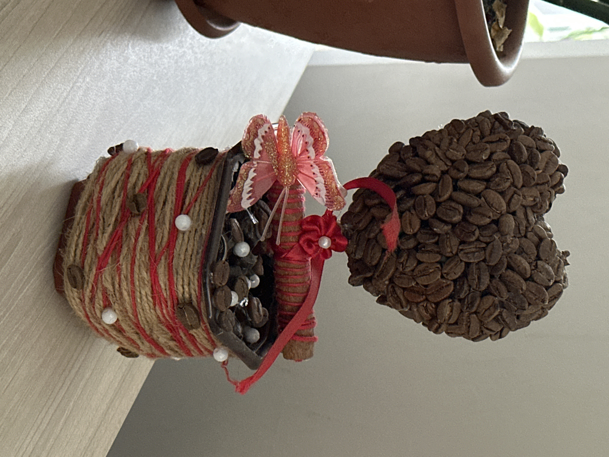 A decorative heart-shaped object made of coffee beans sits in a pot wrapped in twine and red ribbon. A pink glittery butterfly with white accents is attached to the side of the pot. The pot itself appears to be filled with more coffee beans and white beads. The object is placed on a light-colored wooden surface, possibly a shelf or table, next to a terracotta plant pot containing some dry leaves. The background is a plain, light-colored wall. The scene is indoors, near a window that lets in natural light. There is no indication of the city of Hora, Ukraine.