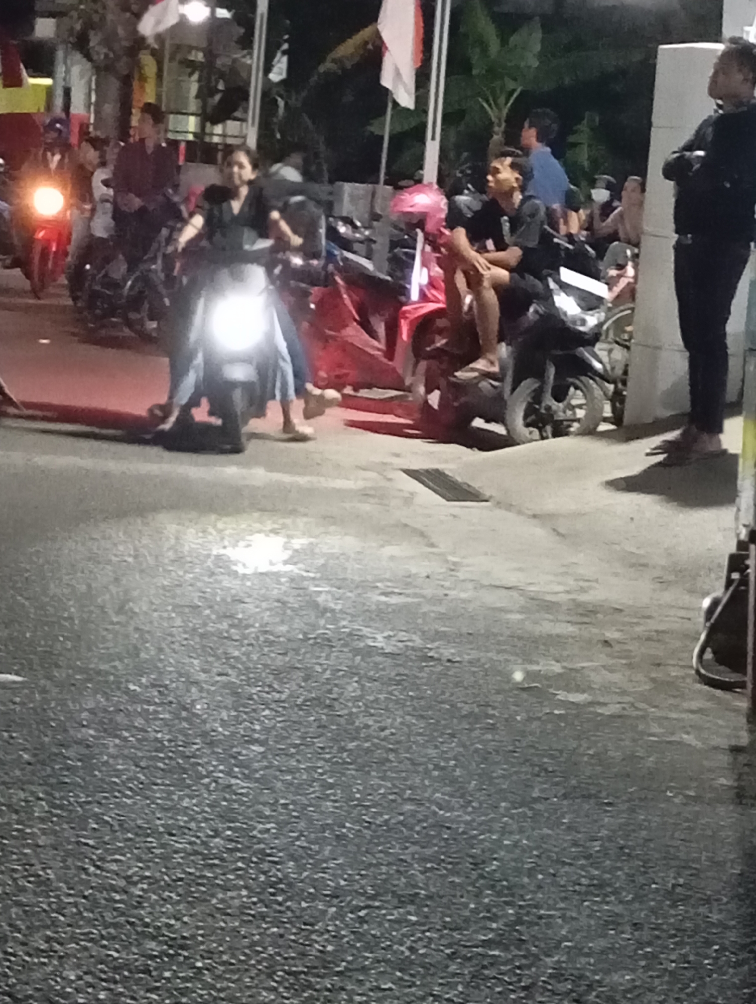 This image depicts a nighttime street scene in Pacitan, Indonesia, likely near a bustling area given the presence of multiple motorcycles and people. In the foreground, a dark asphalt road is visible, wet and reflective, suggesting recent rain or a damp surface. Several motorcycles are parked or in motion.
In the mid-ground, a woman wearing dark clothing is seated on a scooter, its headlight brightly illuminated. To her left, another scooter is partially visible. Further back, a man in dark clothing is seated on a motorcycle, looking to his left. Beside him, a person in a blue shirt stands. Behind them, more individuals and motorcycles are present, with some standing and others seated. The background is dimly lit, with hints of buildings and foliage. A person in dark clothing and pants stands on the right side of the frame, facing away from the viewer. The overall atmosphere is that of a casual gathering or activity taking place on a street at night.