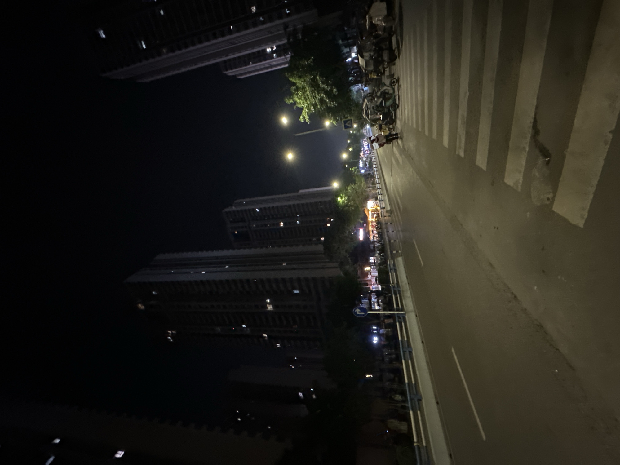 The image depicts a nighttime urban street scene within the Chongqing High-tech Zone, China. Tall, multi-story buildings, predominantly dark with numerous illuminated windows, frame both sides of the asphalt road, suggesting a densely populated area.
In the foreground, a multi-lane road is visible, with white dashed lane markings. A prominent white zebra crossing extends across the right side of the road. A light-colored median or raised curb, lined with a blue and white guardrail, separates the main road from a parallel area to the left, which contains several trees and dimly lit structures or stalls further back.
Streetlights emit a warm, yellowish light, illuminating the road and parts of the pedestrian areas. One particularly bright streetlight is positioned prominently towards the center-right of the image.
Several individuals are present. One person, wearing a light-colored top and dark pants, stands near the right edge of the zebra crossing, facing away from the camera, appearing to interact with a nearby vehicle. A cluster of three-wheeled motor-rickshaw style vehicles, some with canopies, and one blue/green two-wheeled scooter are parked or stationary on the right side of the road near the crossing. Other people are visible in the background, particularly on the left side, near what appears to be commercial establishments with illuminated signs.
Road signs are also visible: a circular blue sign with a white right-turning arrow is on the left side of the road, mounted on a pole, and a square blue sign with a white pedestrian symbol is further down the road, closer to the center, indicating a pedestrian crossing. The sky is dark, typical of a night scene, with a subtle glow near the horizon, indicative of urban light pollution.