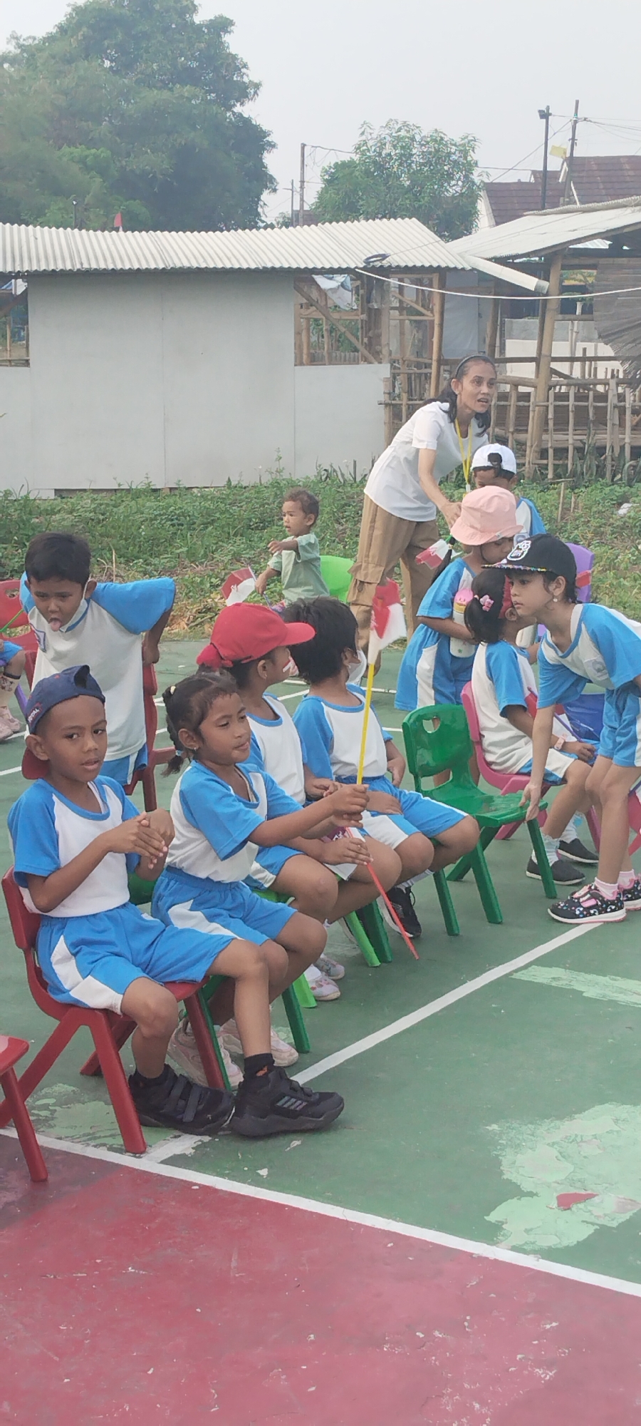 This image depicts a group of children, likely attending a school event in the city of Taban, Indonesia, as evidenced by the Indonesian flags being held by some of the children. The children are seated on colorful plastic chairs arranged on a green and red outdoor court. They are wearing matching blue and white uniforms, with some wearing hats. A woman, presumably a teacher or supervisor, is standing behind the children, observing them. The background shows a fence, vegetation, and a simple building structure, suggesting a less developed area. The overall atmosphere appears to be one of an outdoor gathering or activity for young children.