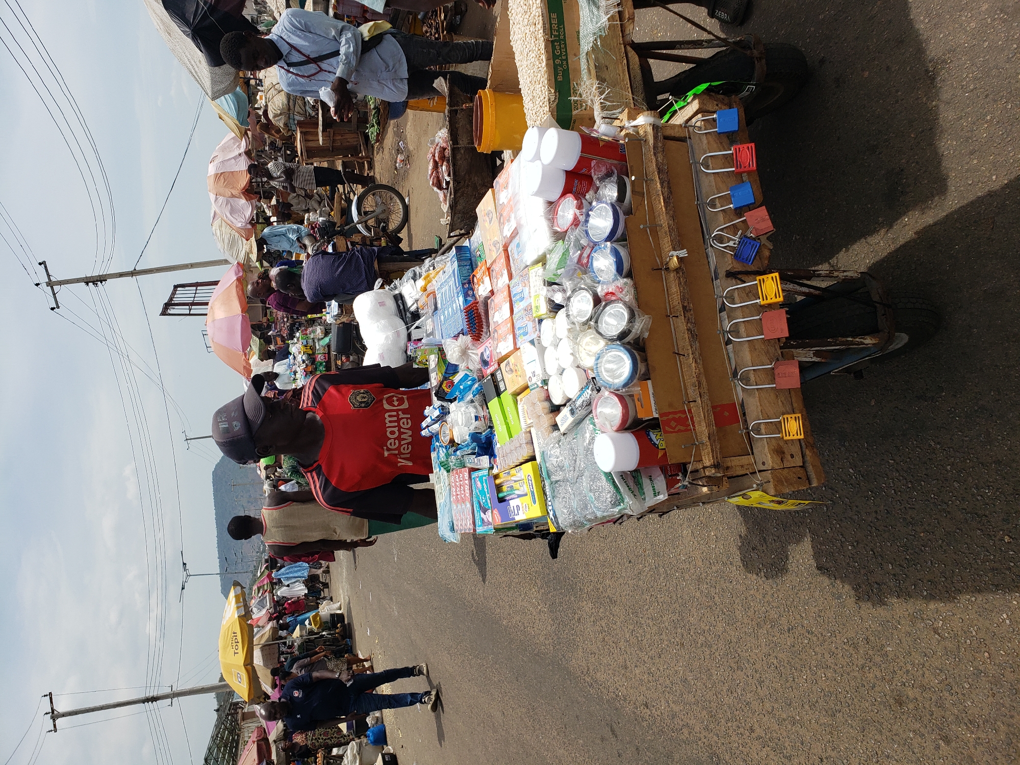 The image depicts a mobile street vendor in Lokoja, Nigeria, selling a variety of household and hardware items from a wooden cart with wheels. The cart is heavily laden with goods including padlocks, flashlights, tape, stationery, and other miscellaneous products. A man wearing a red TeamViewer branded shirt and a cap is positioned near the cart. The vendor's location appears to be along a busy street with other vendors and pedestrians present. Some individuals are under umbrellas, presumably for sun protection. In the background, there are views of surrounding buildings, power lines, and a distant hill or mountain. The ground is paved. A yellow Topit-branded umbrella is visible near the curb of the street.