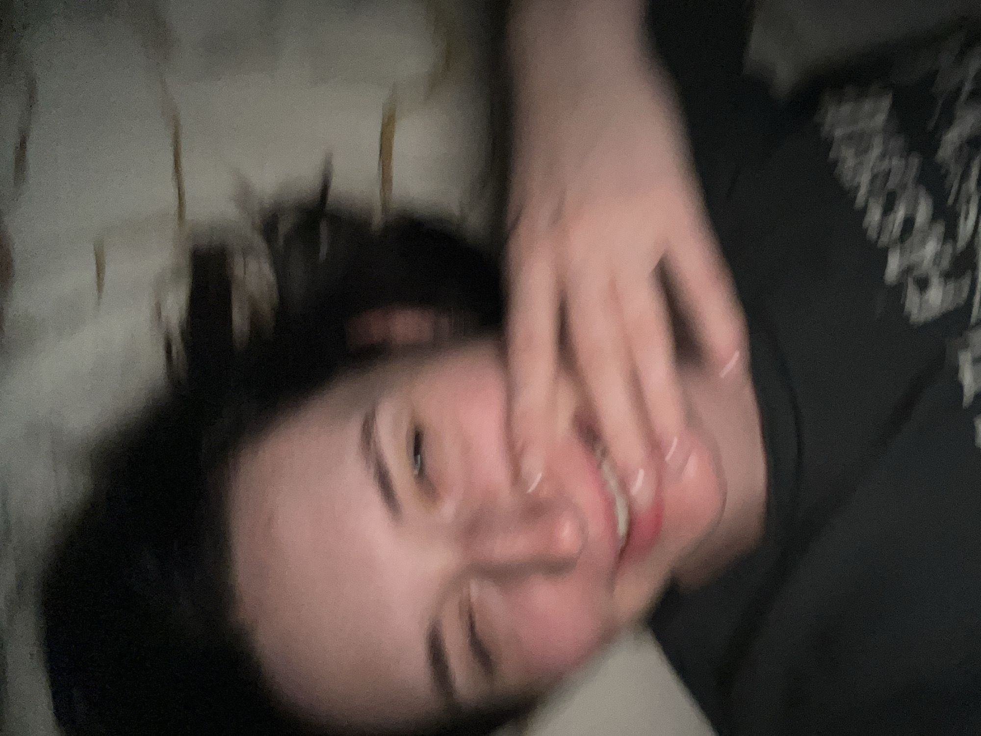 A blurry, close-up photograph depicts a single individual, appearing to be a young adult female, positioned horizontally within the frame. The subject has dark hair and fair skin, with a noticeable flush across her cheeks. She is smiling, revealing both upper and lower teeth, with her mouth slightly open. Her left eye is partially closed, consistent with a winking or squinting expression. Her right hand is raised, partially obscuring the lower right portion of her face and mouth. The fingers of the raised hand are slightly spread, and the fingernails are visible, appearing to have a light or clear polish. She is wearing a dark, likely black, crew-neck t-shirt with indistinct light-colored graphics visible on the lower right quadrant of the garment. The background is uniformly blurry and light-toned, suggesting an indoor setting with no discernible specific details. The prominent motion blur throughout the image indicates either subject movement, camera shake, or low light conditions during capture. The exact location within the city of Tyumen, Russia, cannot be determined from the visual information present in the image.