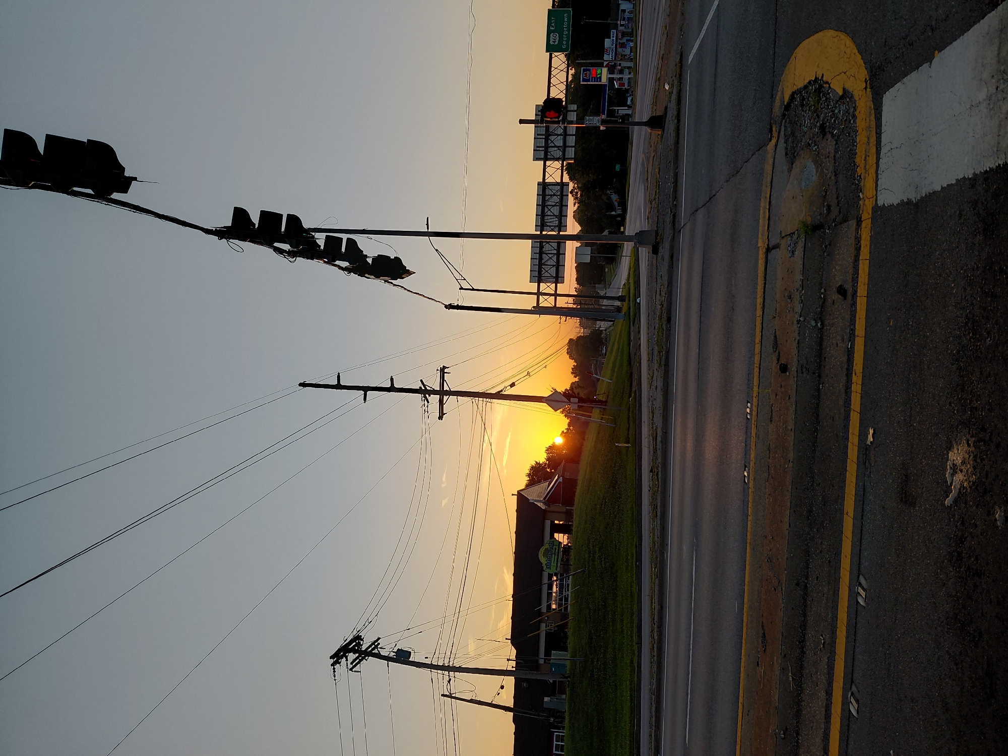 This is a photograph of an intersection at sunrise in Frankfort, Kentucky, United States of America. The sky is a pale orange and yellow, indicating the early morning sun. Several telephone poles and power lines stretch across the sky, silhouetted against the sunrise. In the background, a long, low building with a dark roof sits on a grassy hill. A sign with green lettering can be seen in front of the building. To the right of the frame, a traffic light hangs over the intersection. Further down the road, highway signs indicate "460 East Georgetown." A gas station with a Marathon Food Mart sign is also visible. The foreground shows an asphalt road with yellow lane markings and a crosswalk. The overall scene depicts a quiet, early morning at a suburban intersection.