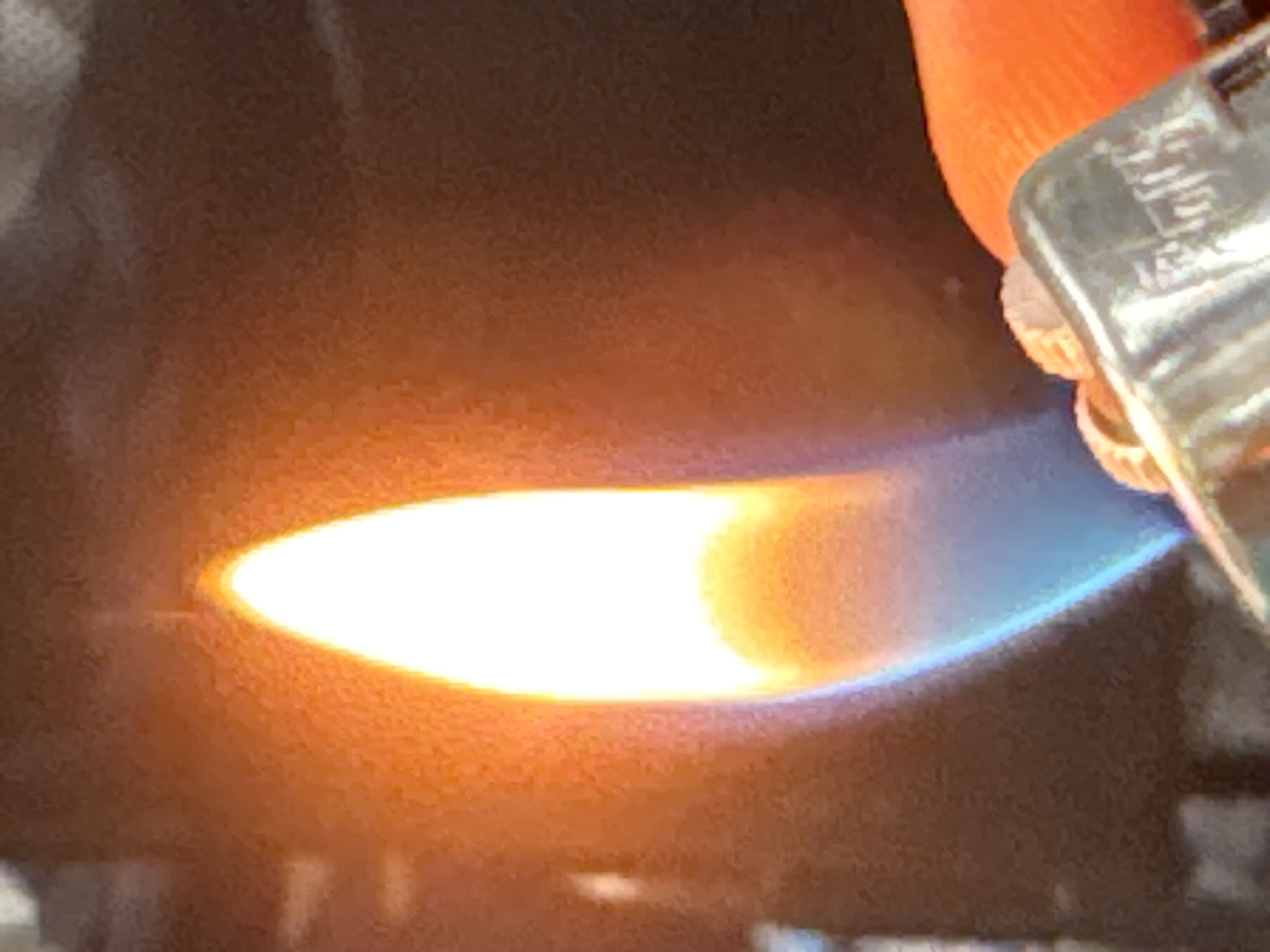 The image shows a close-up of a flame, likely from a lighter, producing a bright, elliptical shape with yellow and blue hues. The flame appears to be the primary subject, emanating from a metal object partially visible on the right side of the frame, which has some engraved text that is illegible due to the focus and lighting. A fingertip, with an orange hue, is also visible at the top right, suggesting someone is holding the object. The background is dark and out of focus, with some textures that could indicate a rough surface or fabric. The lighting is dominated by the intense glow of the flame, casting a warm light on the surrounding elements. There are no clear indicators of time of day, weather, or specific location, although the cropped images provide slight hints of texture that are not definitive. The overall impression is one of focused observation of a small, bright phenomenon, possibly in an indoor setting due to the lack of external light cues.