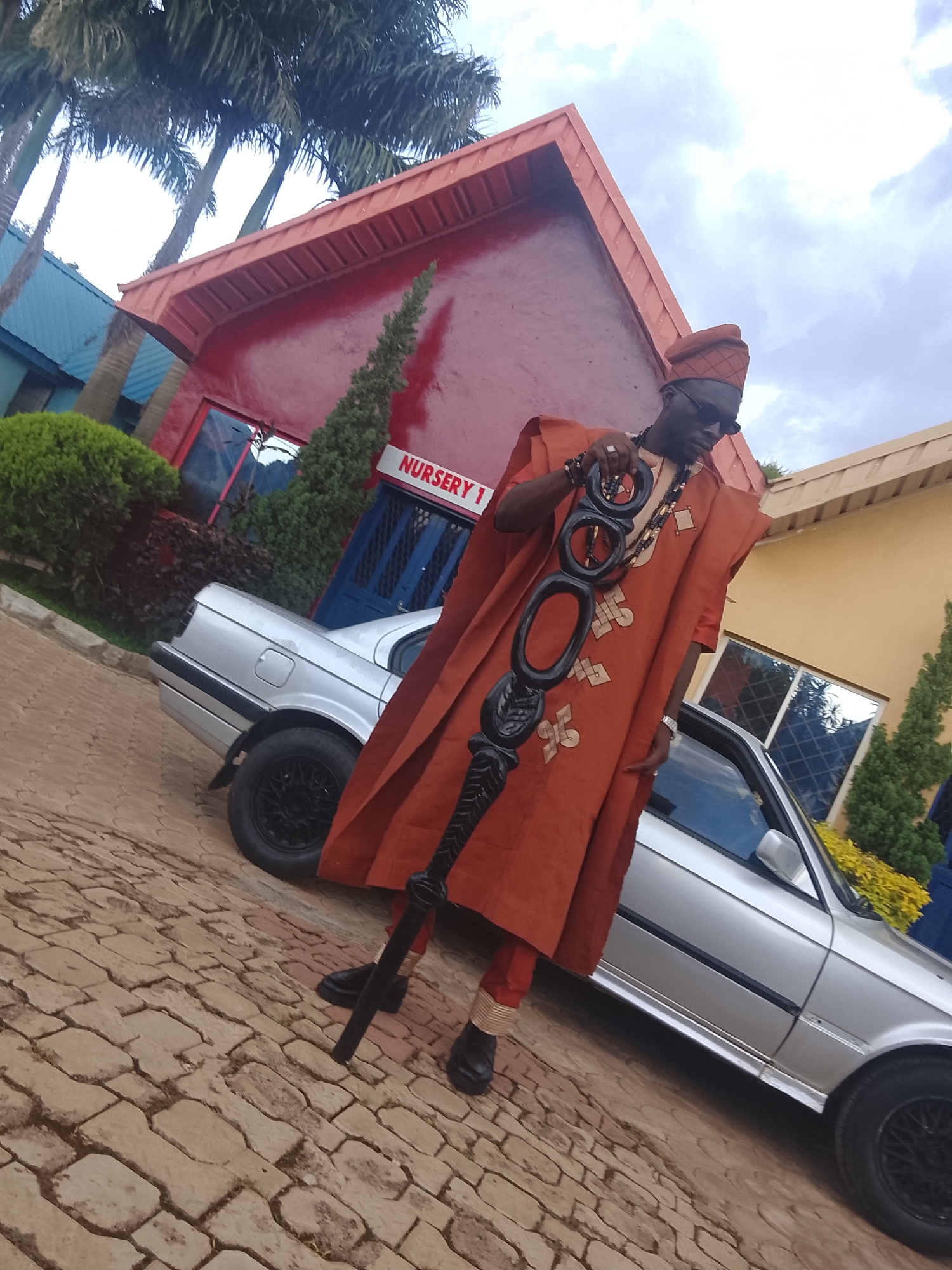 A man dressed in traditional Nigerian attire, specifically an orange agbada and a matching kufi hat, stands in front of a silver sedan. He is wearing dark sunglasses and holding a staff adorned with black, circular carvings. The man is positioned on a paved walkway in front of a building with a red roof and a sign that reads "NURSERY 1." The setting appears to be a public space in Bukuru, Nigeria, with palm trees visible in the background. The overall scene is captured from a low-angle perspective, emphasizing the man and his attire.