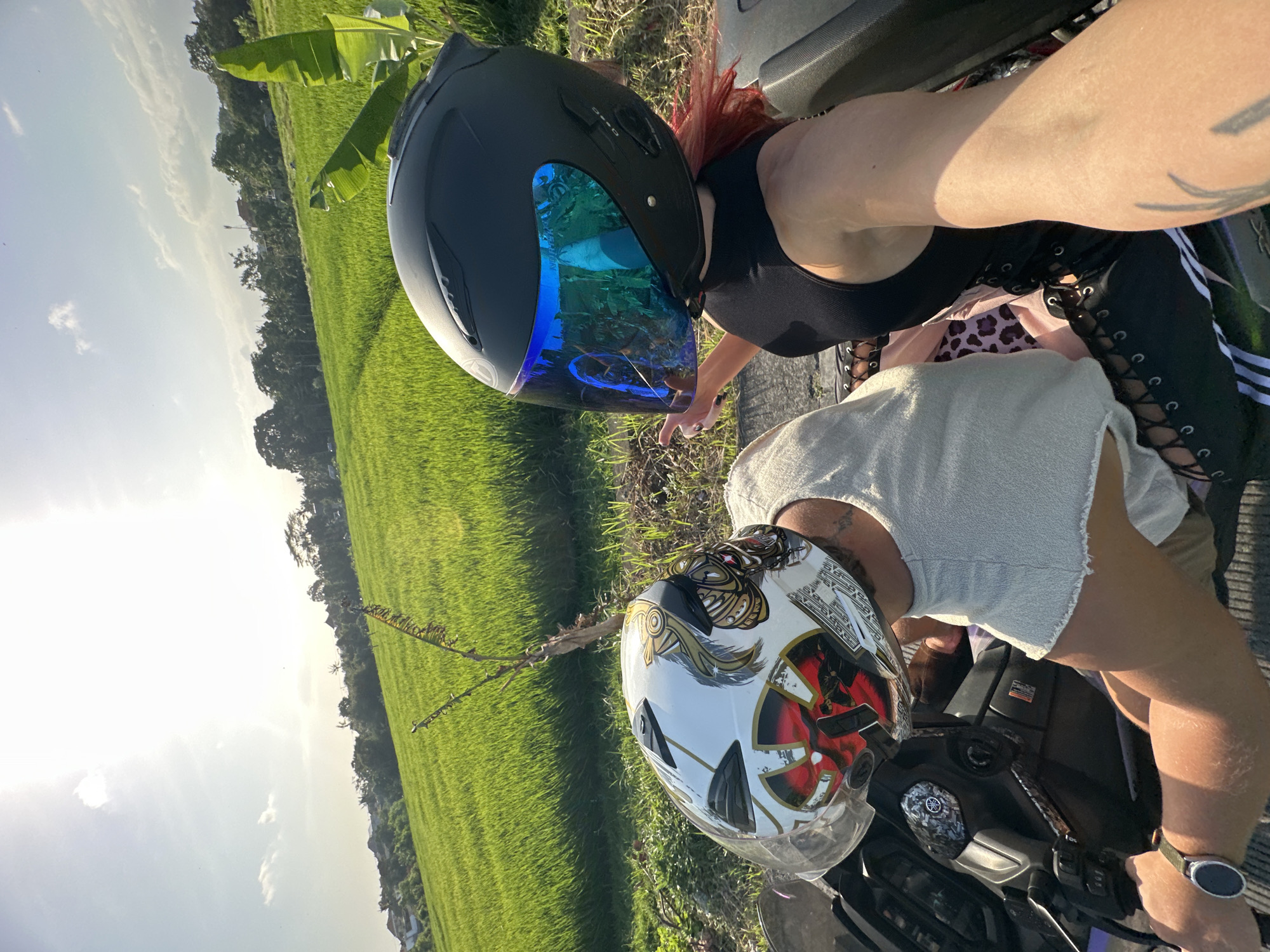 Two individuals wearing motorcycle helmets are seated on a motorcycle, facing forward. The individual on the left wears a white helmet with a red and gold design, and a light-colored top. The individual on the right wears a black helmet with a reflective blue visor and a black sleeveless top. The background features a lush green rice paddy under a bright, sunny sky with scattered clouds. The location is identified as North Kuta, Indonesia. The scene captures a moment of travel or leisure amidst a rural landscape.