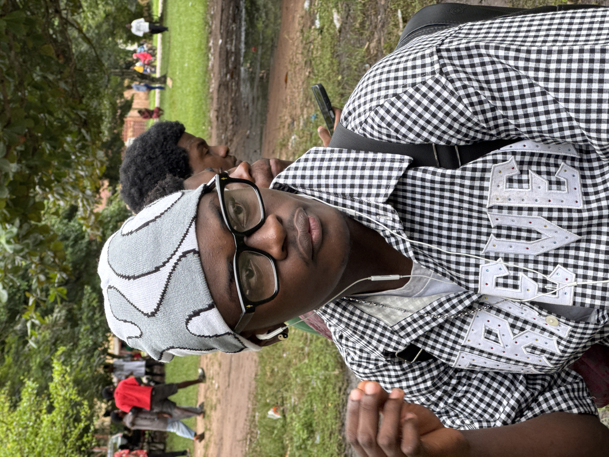 This is a medium shot, taken outdoors, of a young Black man sitting and looking to the right. He is wearing a black and white checkered shirt with "REVE" embroidered on the front in white letters adorned with colorful rhinestones. He also has on a grey and white patterned beanie and black-rimmed glasses. White earbuds are in his ears, and a silver chain necklace is visible around his neck. He is holding a small, reddish object in his left hand. In the background, a second young Black man with curly hair is visible, and further back, other people are scattered in a park-like setting with trees and grass. The ground appears to be a dirt path with puddles of water. The overall impression is casual, and the location suggests a public outdoor space in the city of Ihiagwa, Nigeria.