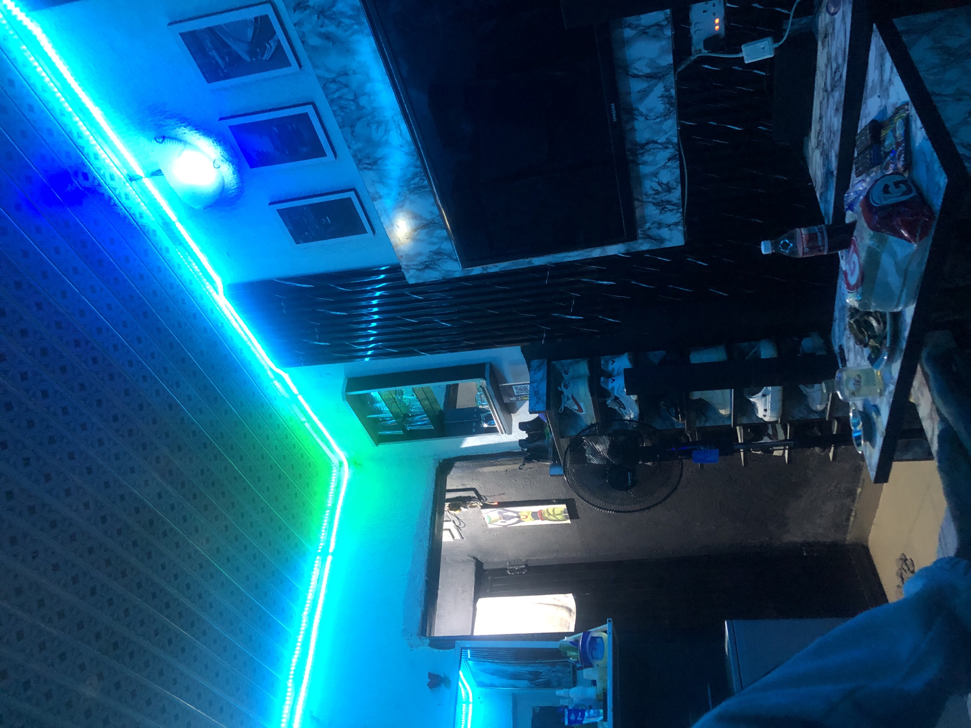 The image depicts an indoor scene, possibly a living room or bedroom, characterized by vibrant blue and green LED strip lighting illuminating the ceiling and walls. A dark-colored ceiling with a patterned texture is visible. On the wall, there are several framed pictures. To the right, a television is mounted on a wall with a marble-like finish. Below the television, a small table holds a bottle of soda, a pack of bread, snacks, and remote controls. To the left, a doorway leads to another area, with a white towel hanging inside. A fan is positioned in the foreground, and shelves filled with shoes are visible behind it. A person's arm wearing a light-colored sweatshirt is partially visible in the lower left corner. The overall ambiance is dimly lit, with the LED lights creating a colorful and modern aesthetic. The location is identified as Unknown, Unknown.