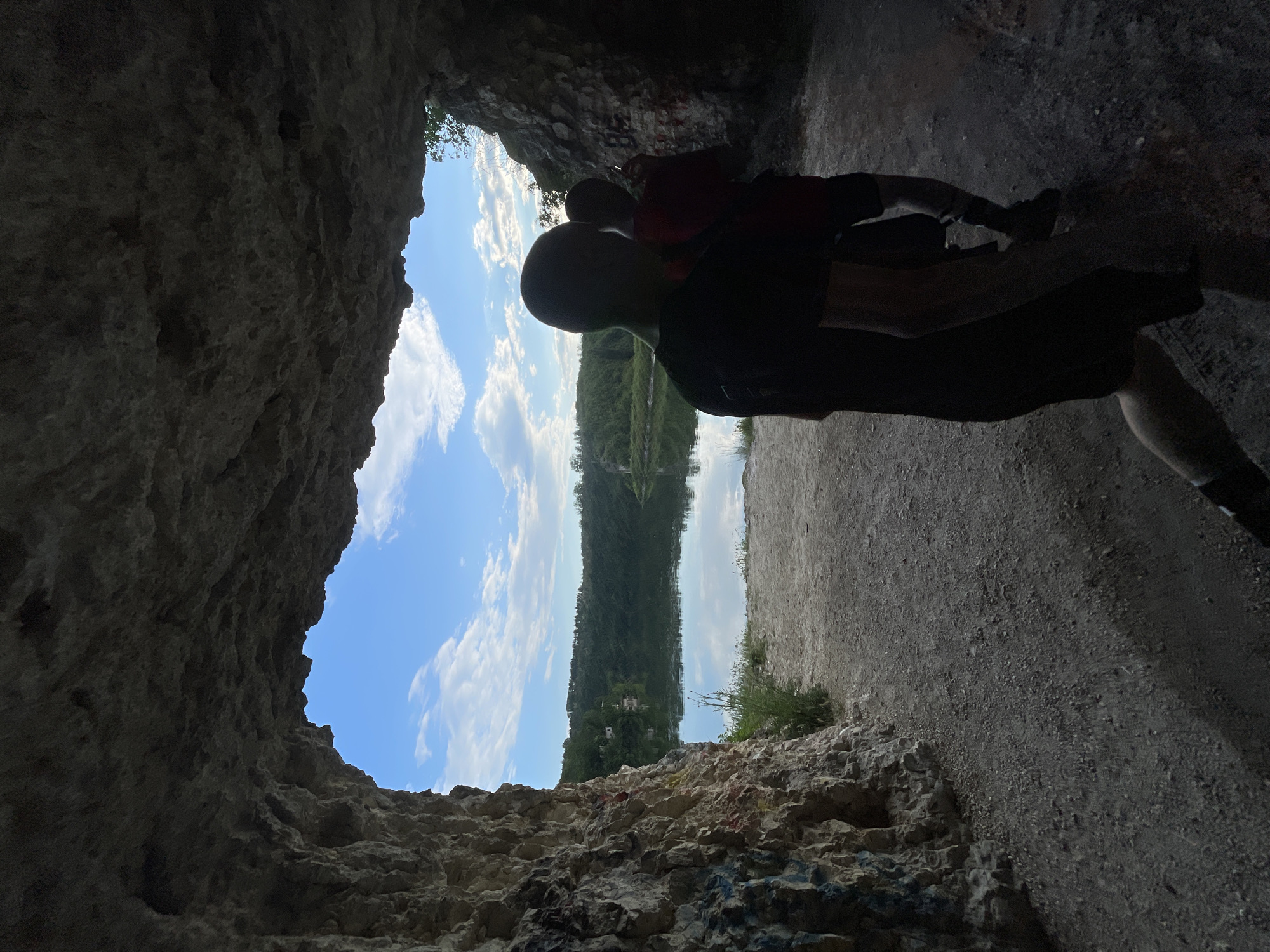A person is silhouetted in the foreground of a cave opening, looking out toward a view of water, trees, and sky. The sky is blue with some clouds. The viewer's perspective is from inside the cave, with the rough, textured rock walls of the cave surrounding the view. A gravelly path is visible to the right of the cave opening. The location is Unknown, Unknown.
