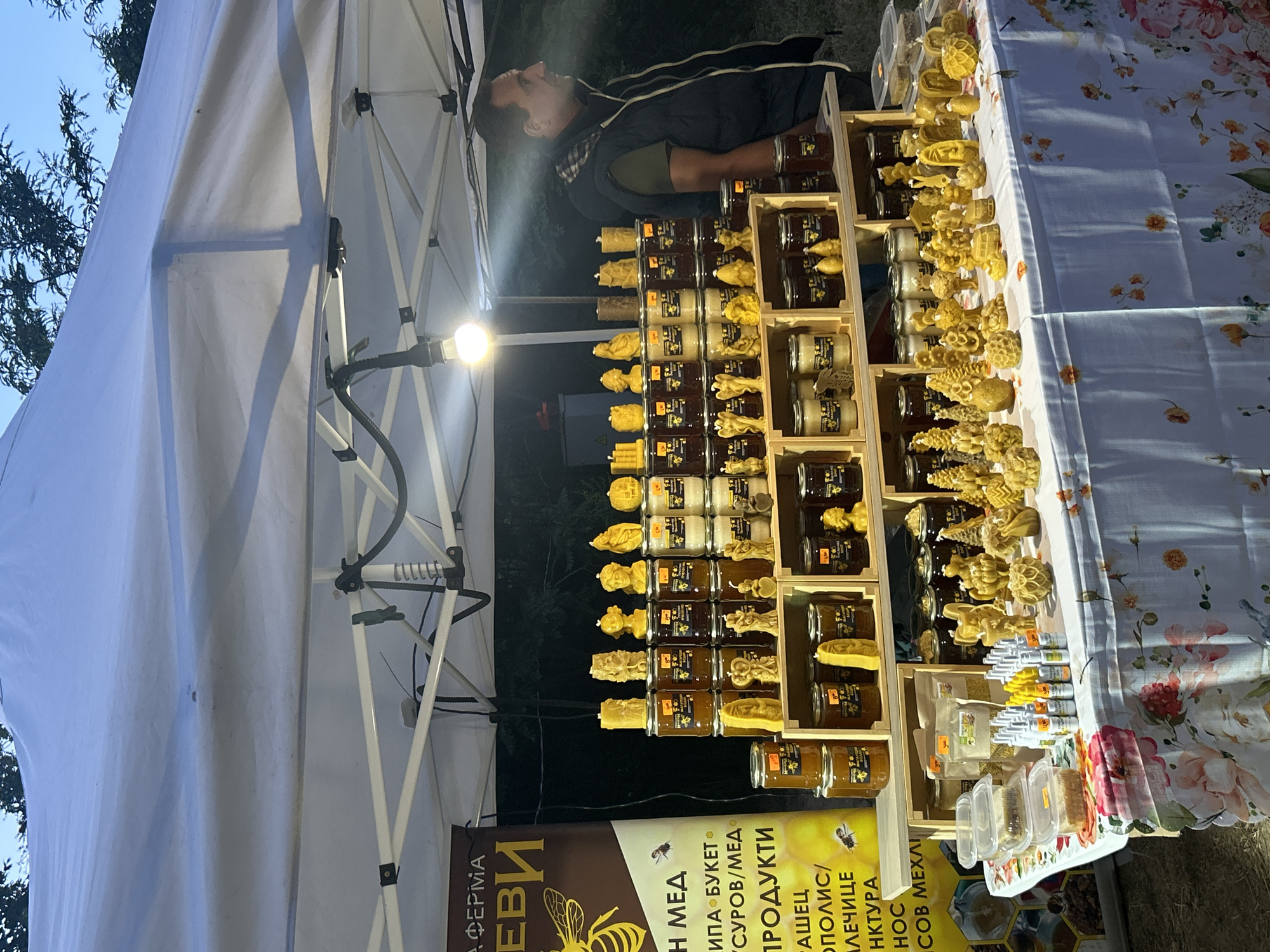 An outdoor market stall displays an assortment of honey products and beeswax candles. The stall is covered by a white canopy, illuminated by a single bulb. On the left, a banner advertises "ФЕРМА" and "МЕД," featuring a graphic of a bee. The stall's shelves are laden with jars of honey and various shaped beeswax candles, including figures of angels, busts, and honeycomb shapes. A man in a dark vest stands to the right of the stall, observing. The table is draped with a floral-patterned cloth.