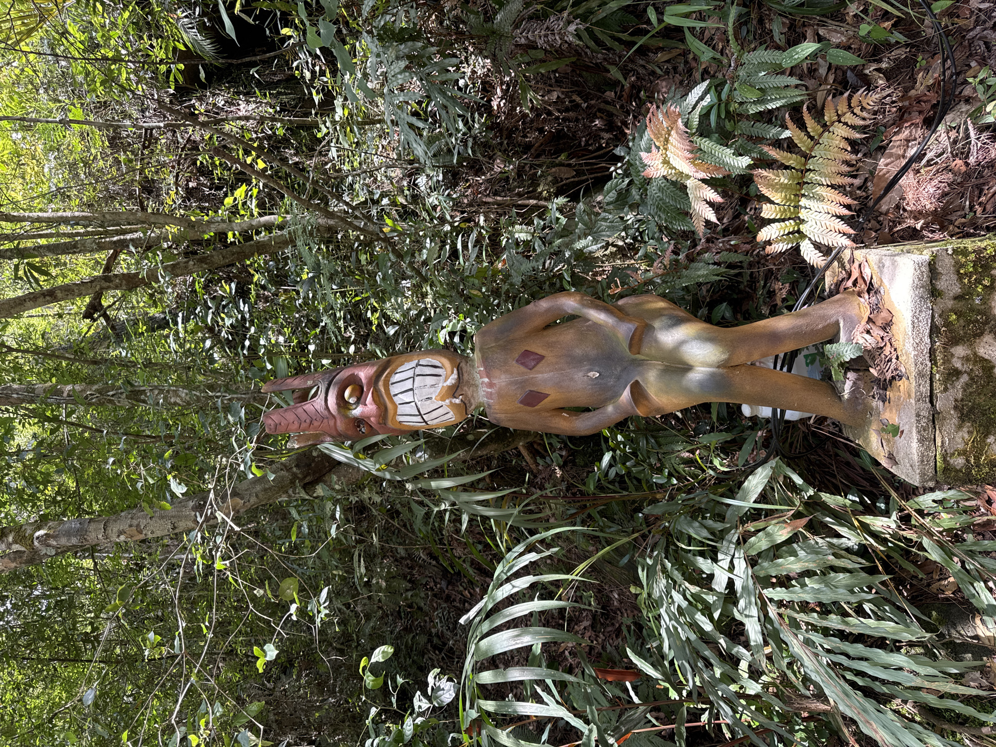 The image shows a wooden statue with a humanoid form and exaggerated facial features, resembling a tiki figure, standing in a lush, overgrown area. The statue is light brown with darker accents on the face and body. It has large, square teeth, prominent eyes, and elongated ears. Brown diamond-shaped figures are also painted on the body. The statue is positioned on a concrete base covered with moss. The surrounding environment includes a variety of plants, ferns, and trees. The foliage suggests a natural, possibly jungle-like setting. The location is Lang Biang, Da Lat, Vietnam, as specified in the prompt.