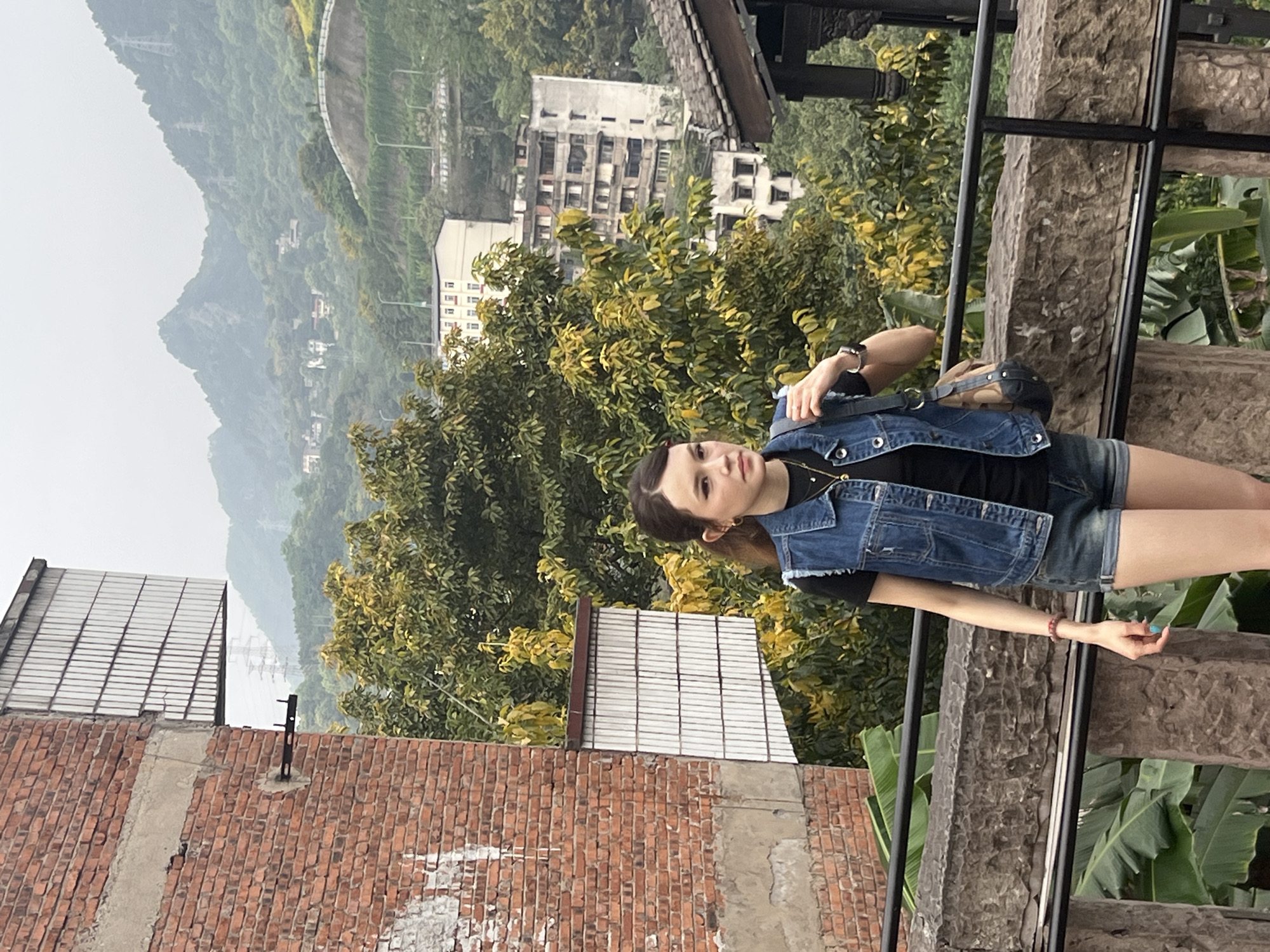A young woman stands in the foreground, wearing a black t-shirt, a blue denim vest, and denim shorts. She has a shoulder bag. She is on a stone staircase, with a black metal railing on the right side of the frame. The background features lush green trees, buildings, and a hazy mountain landscape. A brick building is visible in the lower left corner. The photo suggests an outdoor setting in the Nan'an District of China.