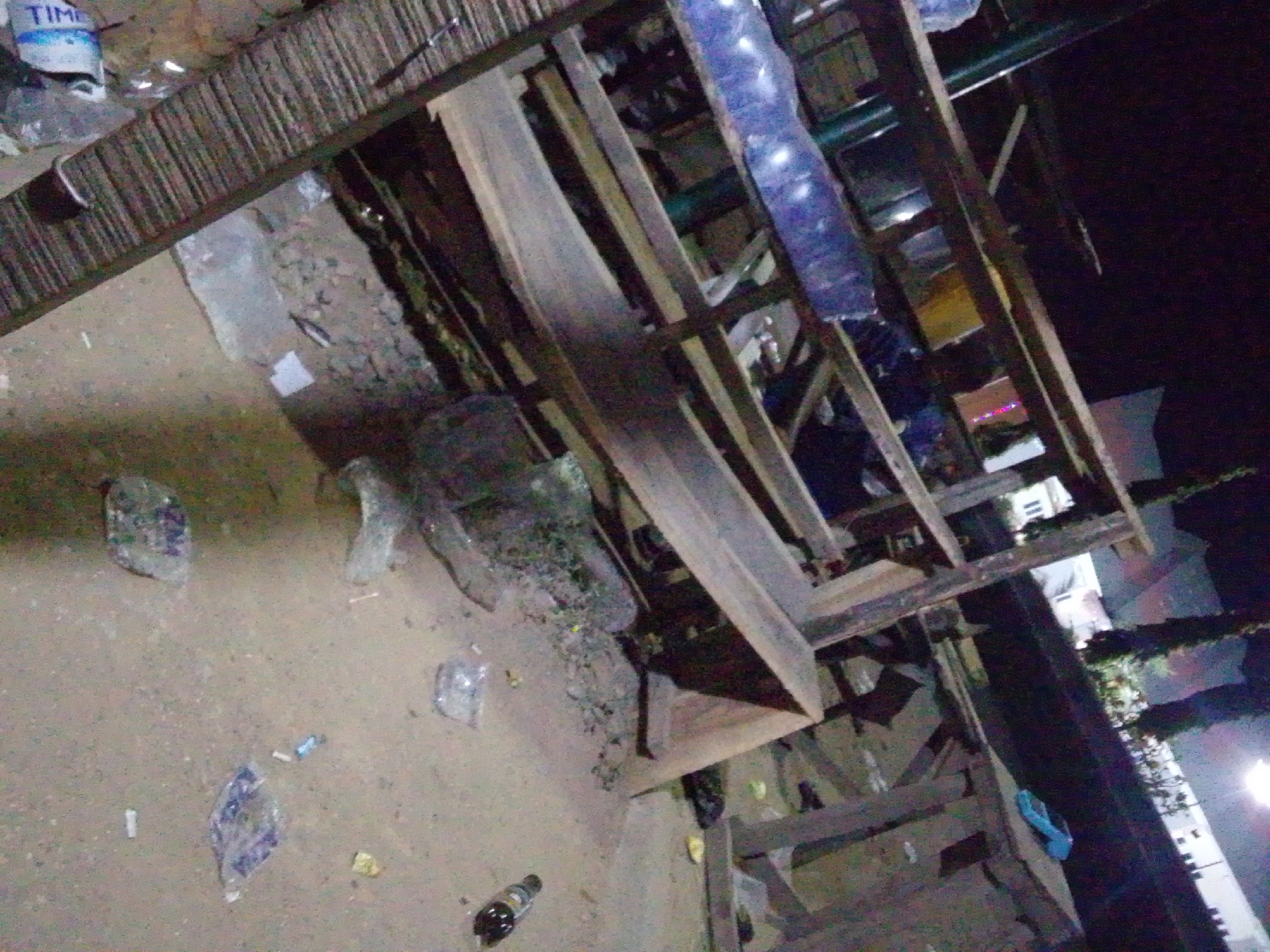 The image shows an outdoor scene in what appears to be an urban environment, potentially Ajaokuta, Nigeria. The scene is littered with plastic bags and bottles, suggesting an area with some degree of waste disposal issues. A wooden structure, possibly scaffolding or part of a temporary building, is prominently featured. The lighting is dim, indicating a nighttime or low-light environment. Buildings are visible in the background, partially obscured by darkness and foliage. There are no people directly visible in the image. The scene is generally unkempt and suggests an area with lower maintenance and potentially lower socioeconomic status. A bottle and a can of "TIME" brand is visible on the left side of the photo.