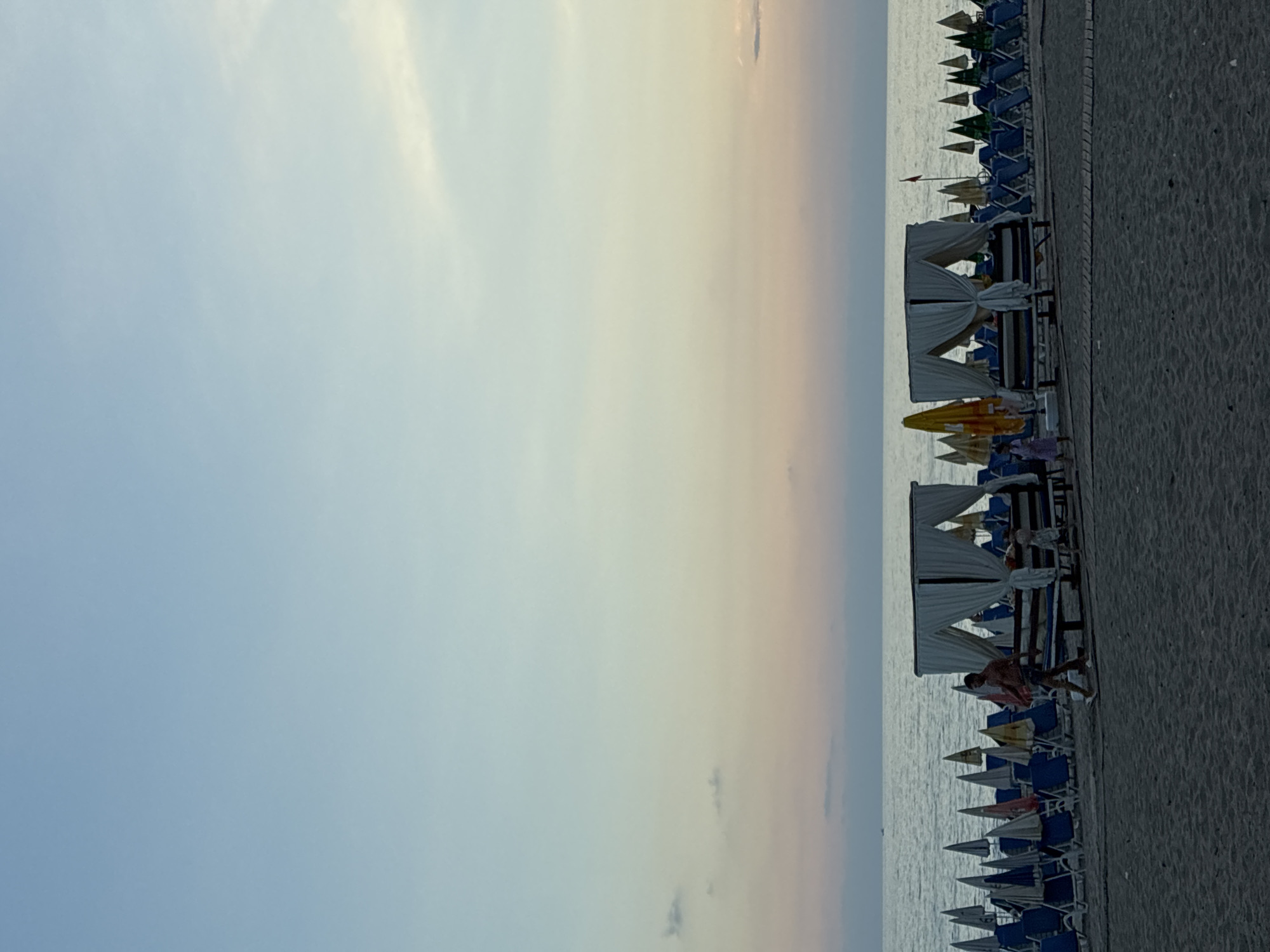 The image depicts a beach scene in Alanya, Turkey, likely during sunset or dusk, as indicated by the soft hues in the sky. In the foreground, there is dark sand. A wooden walkway runs parallel to the water. Beyond the walkway, rows of empty blue beach chairs and umbrellas are visible. Two cabanas, draped with white fabric, are positioned on the sand. A man in shorts is walking from left to right in front of the cabanas. A child in a light-colored dress is walking towards the cabanas. The sea is calm and stretches to the horizon, where it meets a sky transitioning from pale blue to soft pink and orange. A few scattered clouds are present in the sky. The overall atmosphere is tranquil.