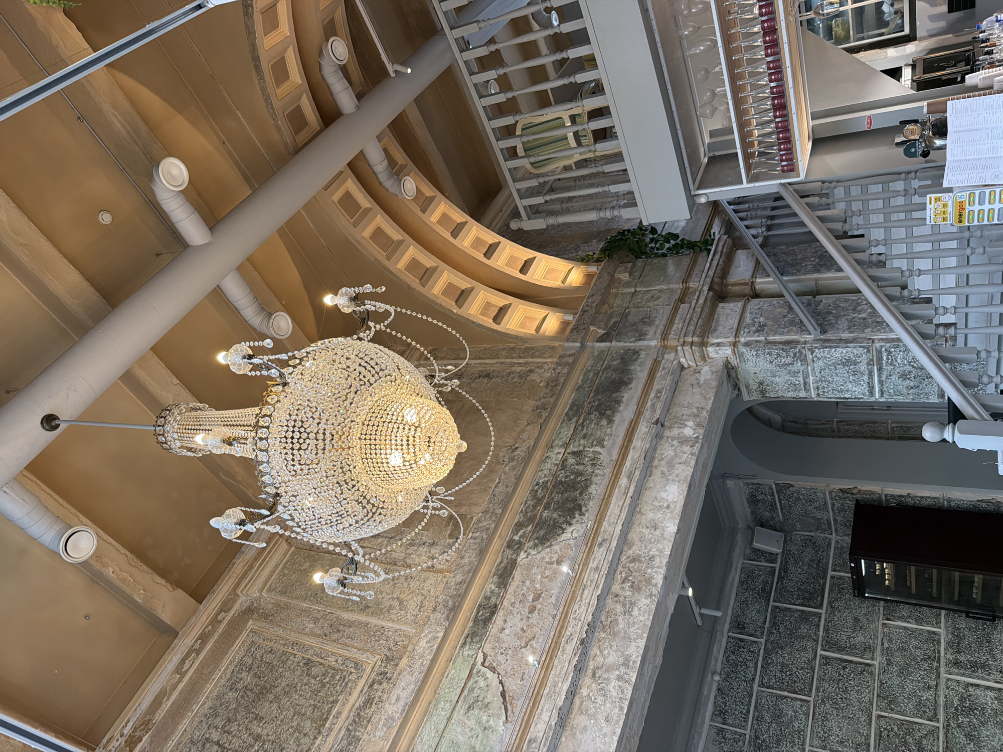 The image shows an interior with a mix of old and modern elements. A large, ornate chandelier is the focal point, hanging from a textured, beige-colored ceiling with decorative molding. A gray support beam runs diagonally across the ceiling, with several round, white ventilation units attached. To the right of the chandelier, the wall appears to be made of stacked gray stone blocks. A staircase with white railings and a glass case holding wine bottles can also be seen in the background. The location appears to be a building in Перовское сельское поселение, Ukraine.